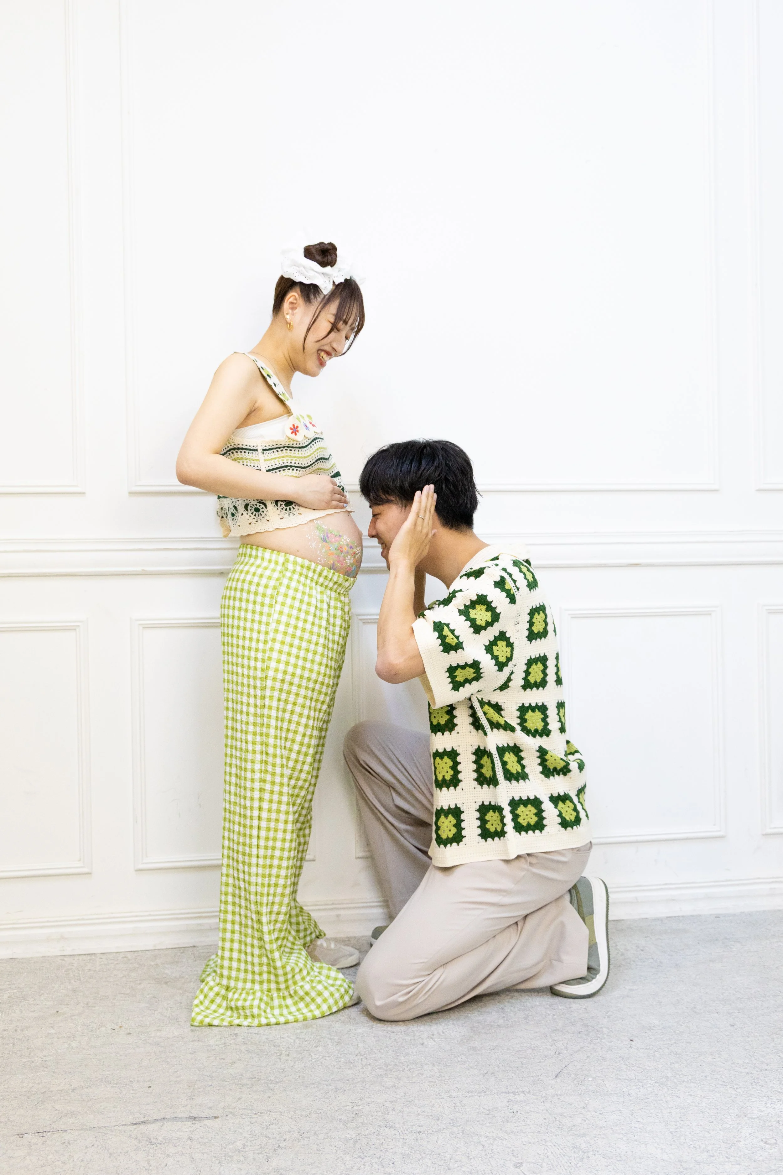 A man kneeling and holding the pregnant belly of a woman smiling, both facing each other in a white room.