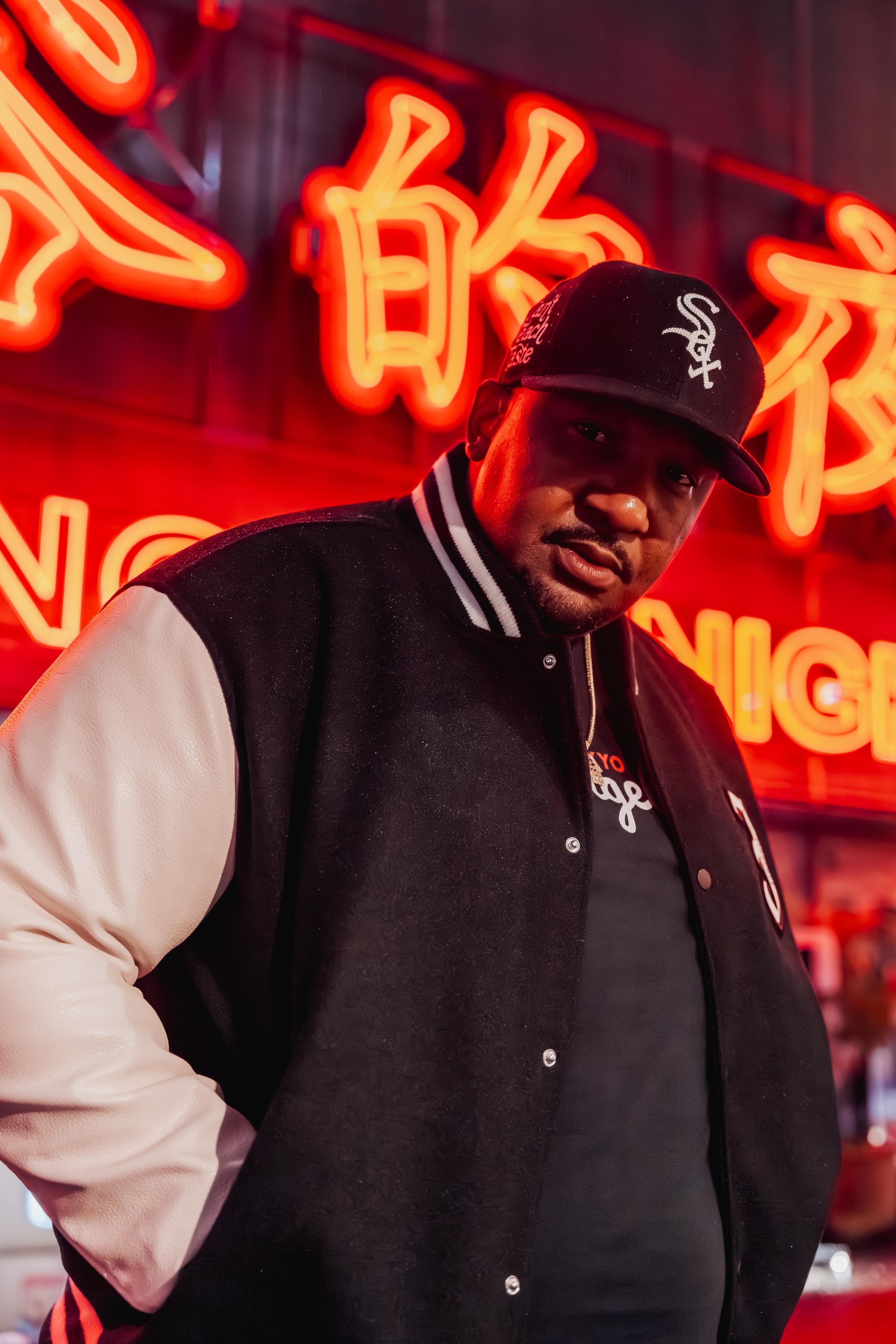 A man wearing a black baseball cap and jacket with white sleeves standing in front of a neon red sign with Chinese characters and English words.