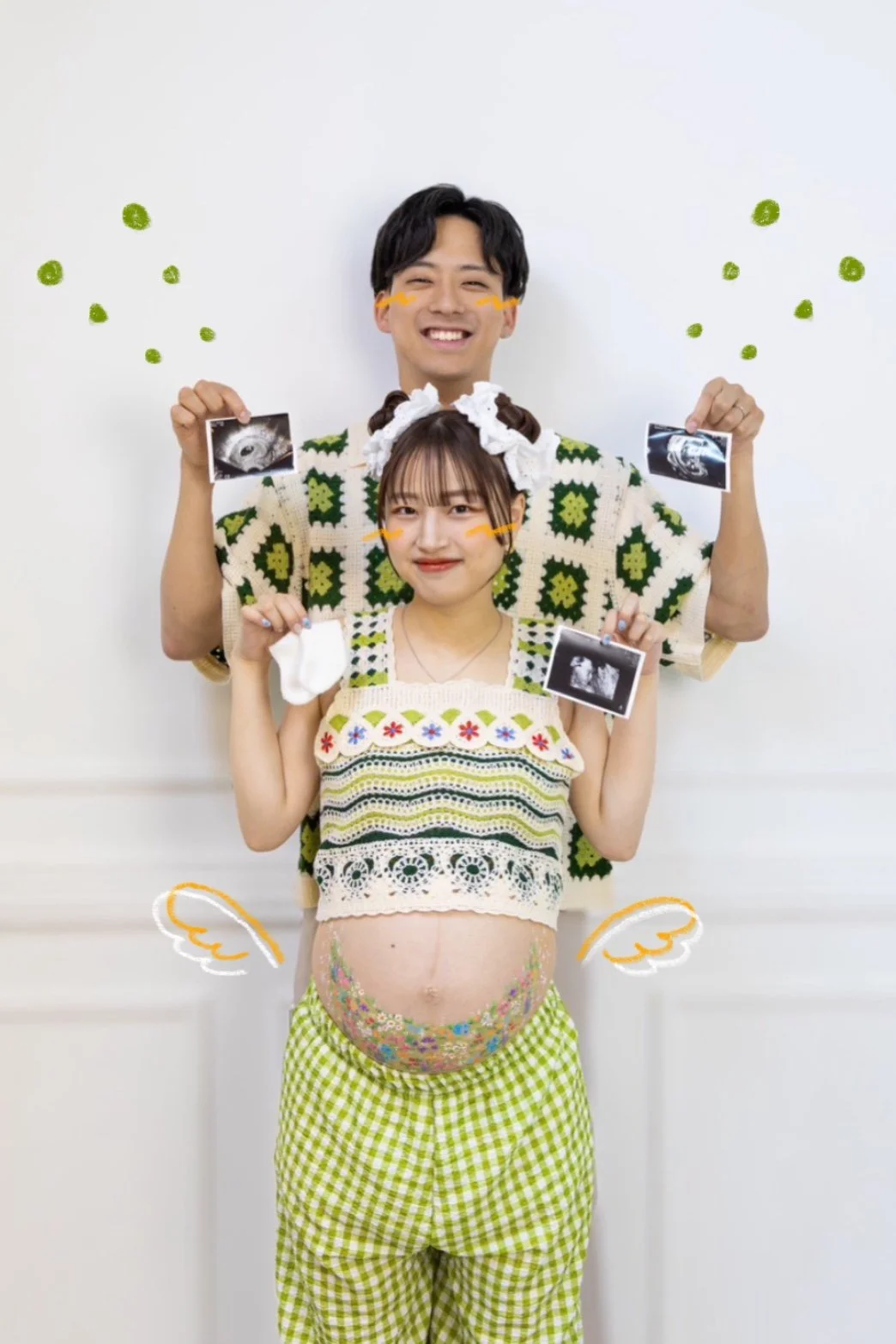 A pregnant woman standing in front of a man, both smiling, holding ultrasound images, with digitally added angel wings and orange lines on their cheeks, and the woman is dressed in a crochet top and green checkered pants.