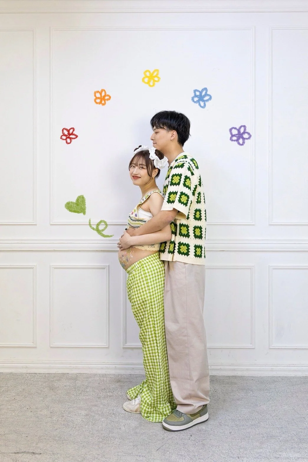 A pregnant woman and a man standing against a white wall decorated with colorful flower and heart drawings. The woman is smiling, wearing a sleeveless top with a gingham pattern and green checkered pants, with her belly painted. The man is standing b