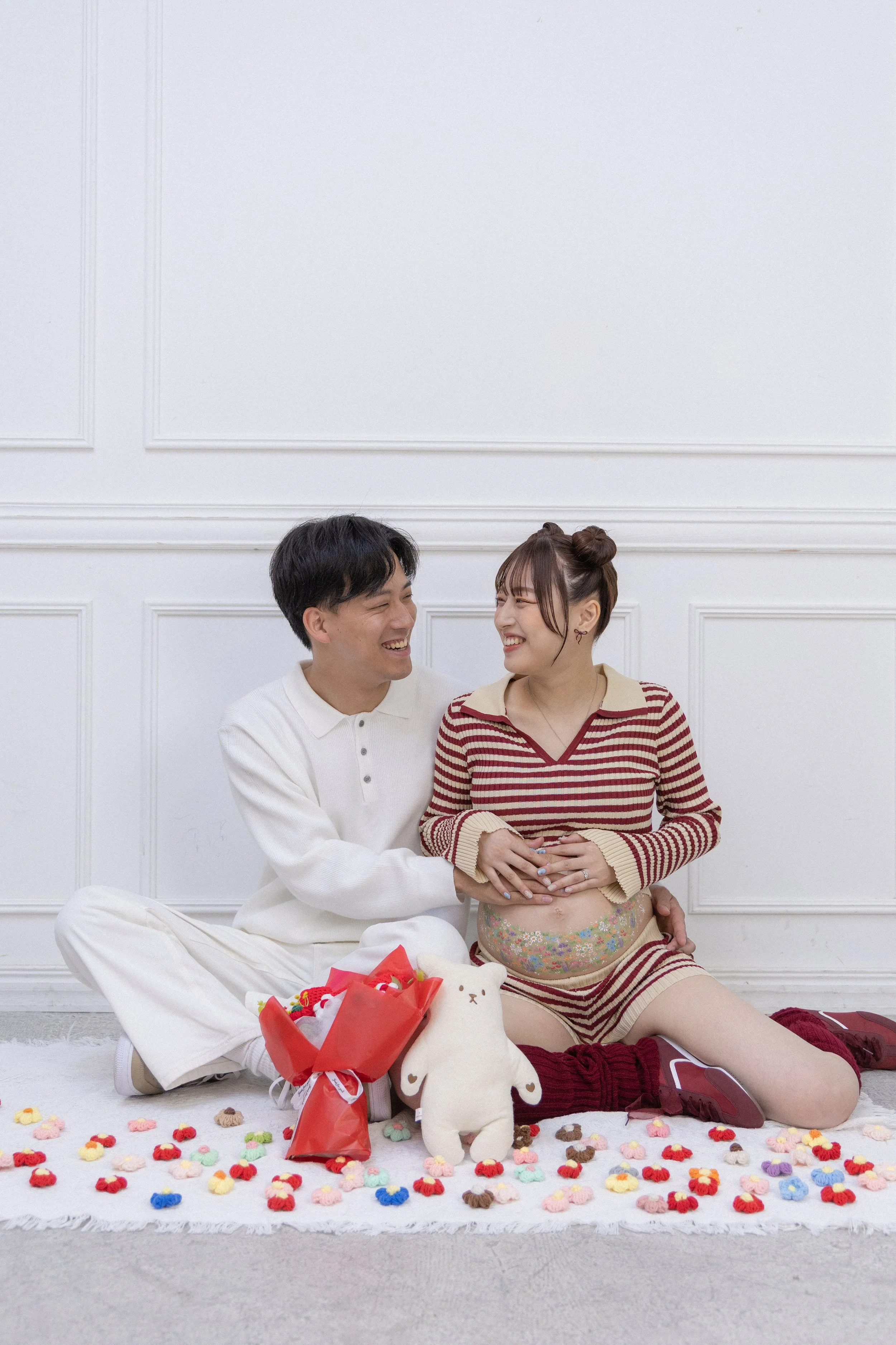 A smiling couple sitting on a white rug with colorful small plush toys and stuffed animals, including a white bear, in a bright room with white walls. The woman, dressed in a striped sweater and shorts, is pregnant and touching her belly while holdin