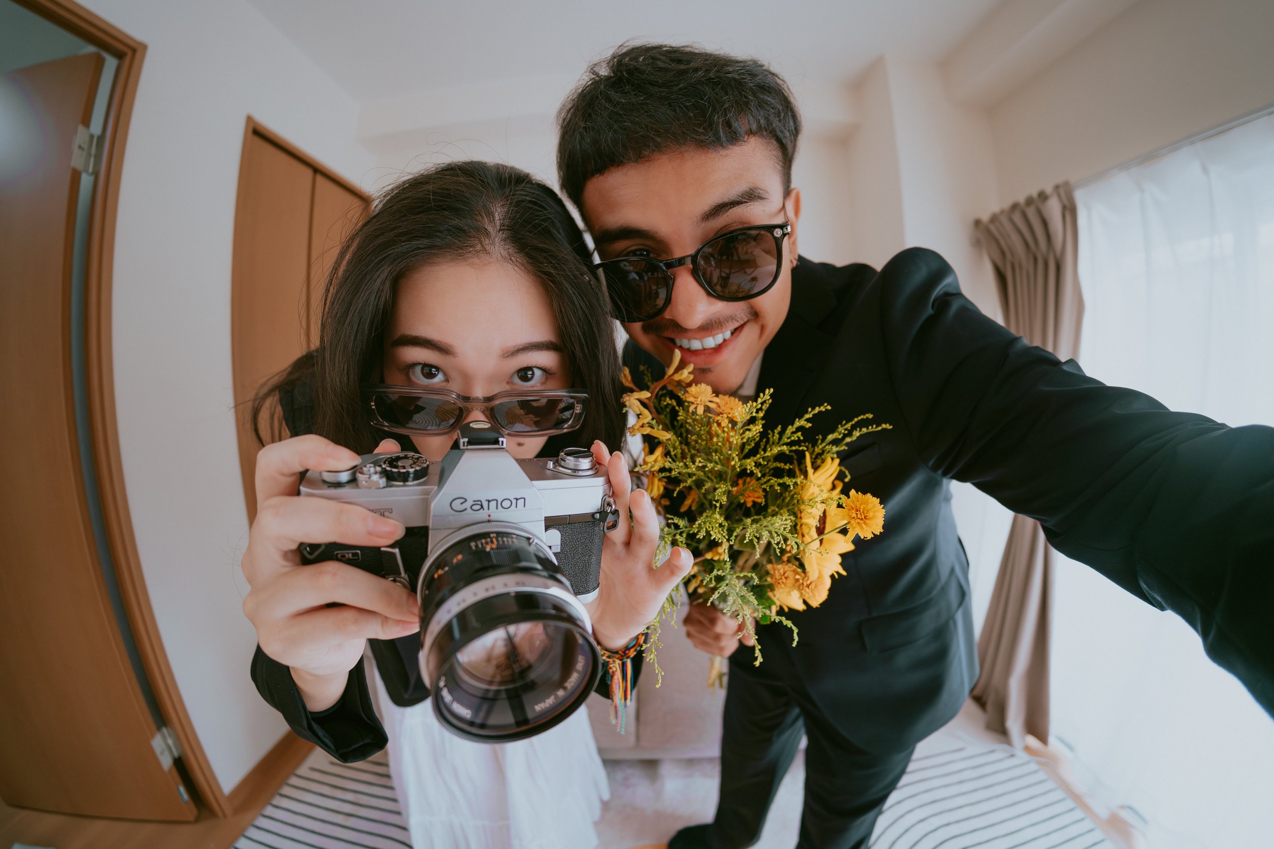 Two young adults taking a selfie indoors. The woman is holding a vintage film camera and wears sunglasses, while the man, wearing sunglasses and a black suit, holds a bouquet of yellow flowers. They are smiling and appear excited.