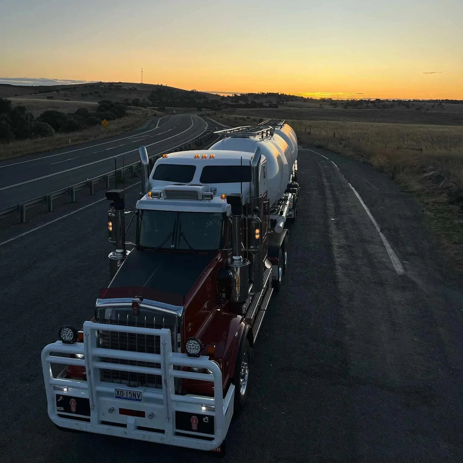 A red fire truck driving on a rural road at sunset with rolling hills and fields in the background.