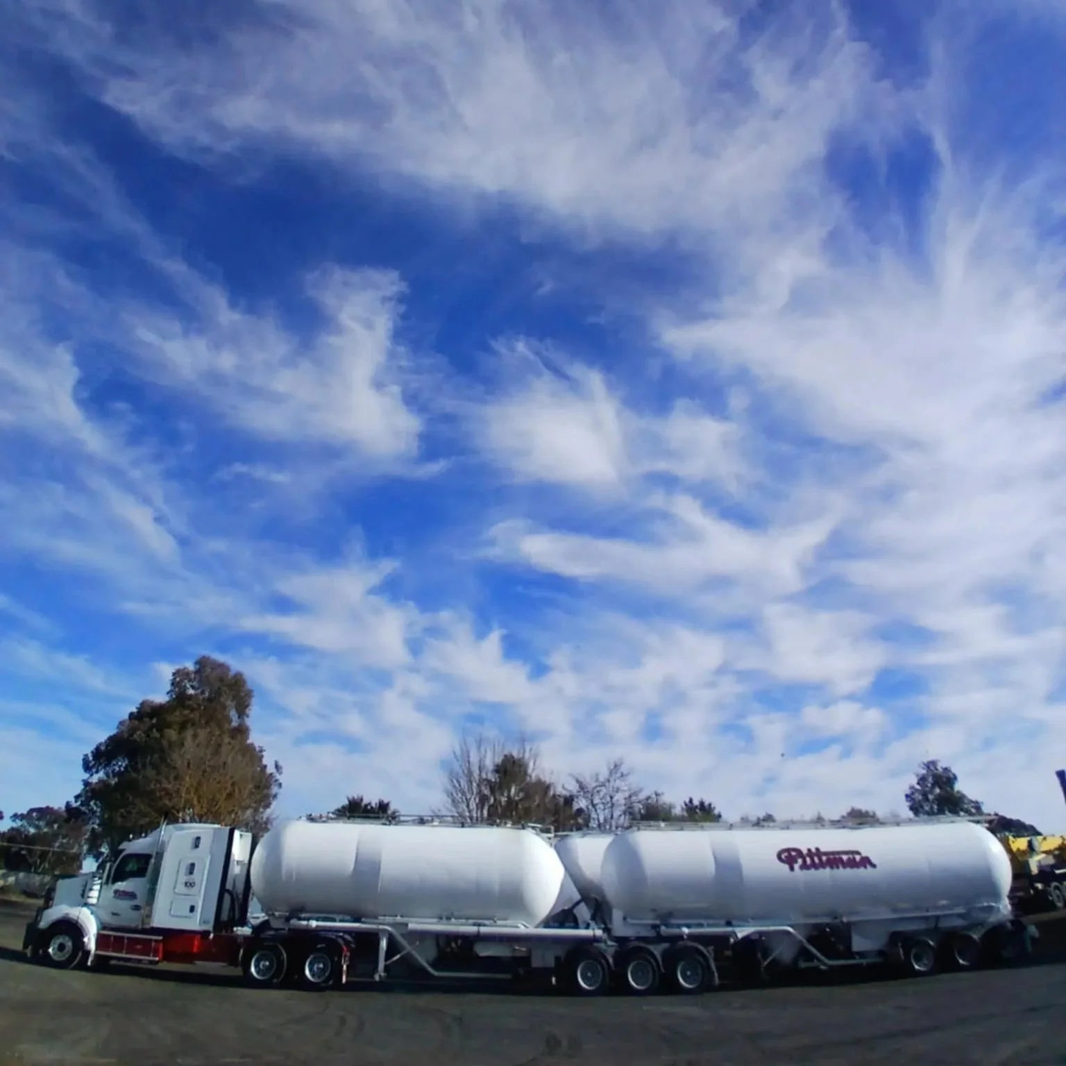 A white tanker truck on the road with trees and a partly cloudy sky in the background.