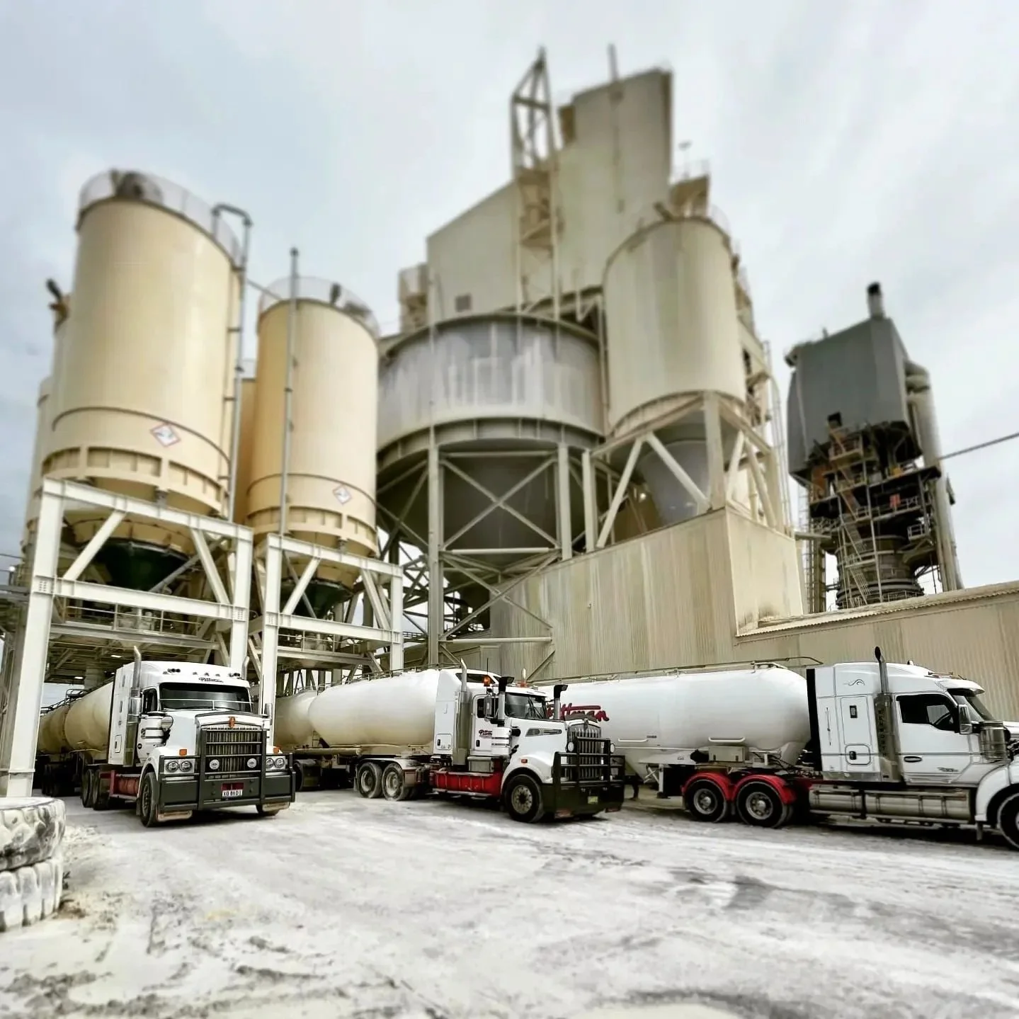Industrial plant with large beige and gray storage tanks, scaffolding, and multiple white and silver tanker trucks parked on a gravel lot under a cloudy sky.