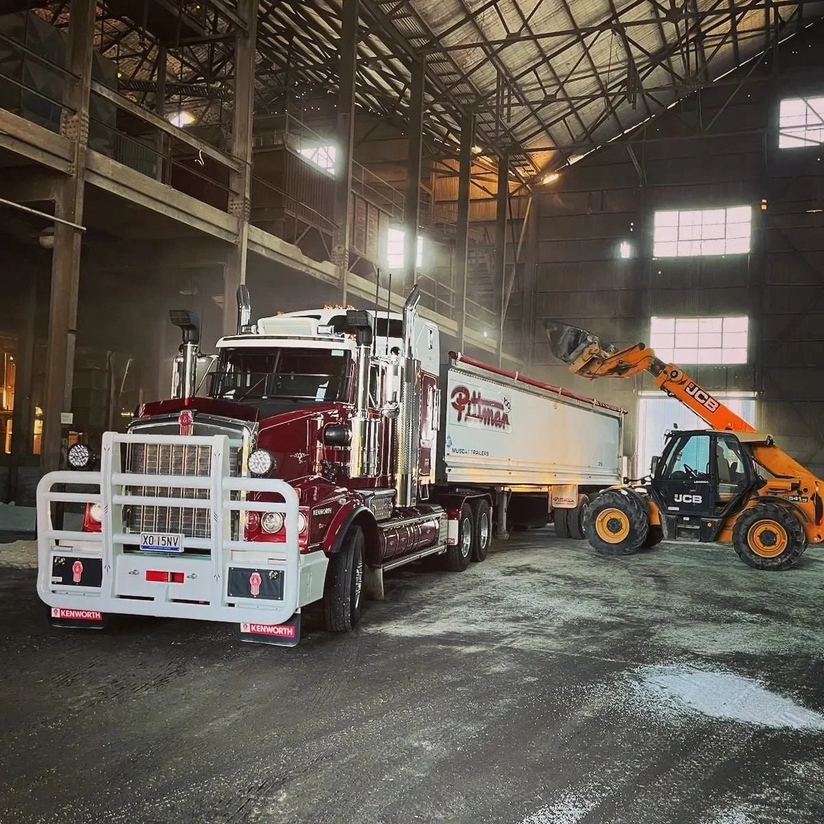 A large Kenworth semi-truck with a white and red cab parked inside a spacious industrial warehouse. An orange JCB telescopic handler is raising a container or equipment. The warehouse has high ceilings, large windows, and metal structures, with some sunlight coming through the windows.