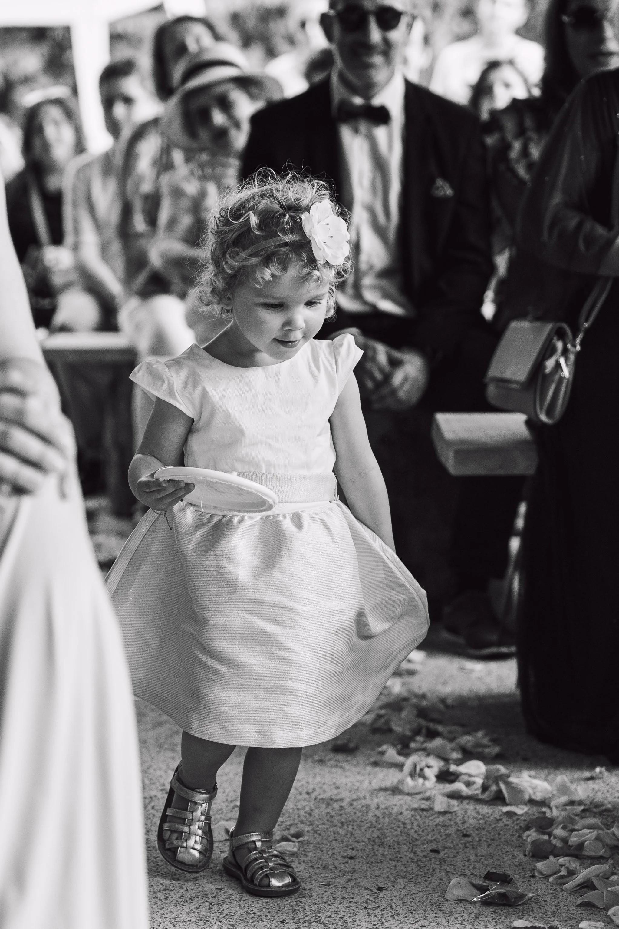 Une petite fille avec une robe blanche, portant une fleur dans les cheveux, marche dans une cérémonie avec des invités en arrière-plan.