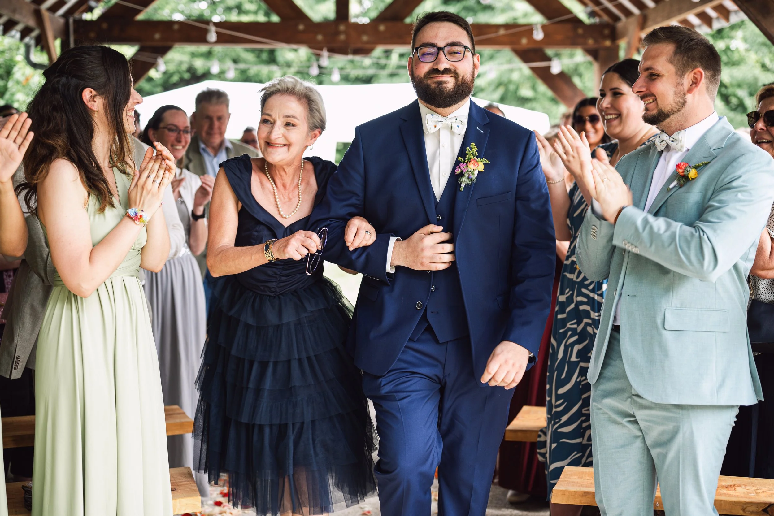 Un groupe de personnes lors d'un mariage en plein air, avec un homme en costume bleu avec un nœud papillon, entouré de personnes applaudissant et souriant.