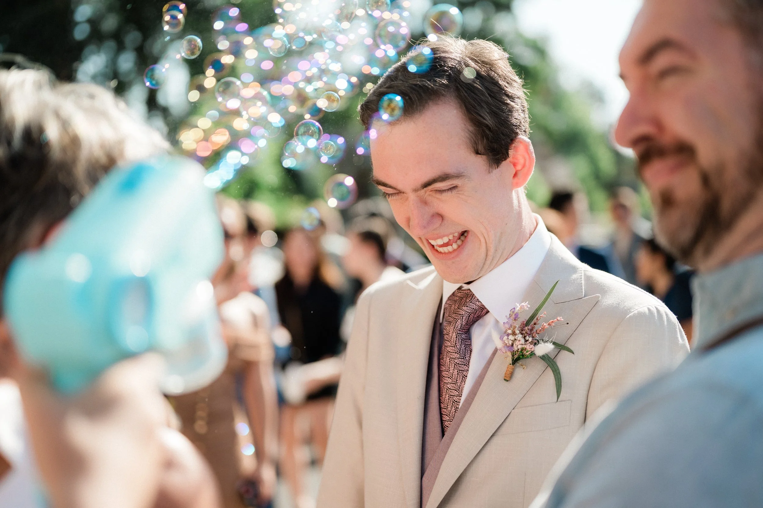 Un homme en costume beige, avec une boutonnière de fleurs, sourit joyeusement lors d'un mariage en plein air, entouré d'invités et de bulles de savon qui flottent dans l'air.