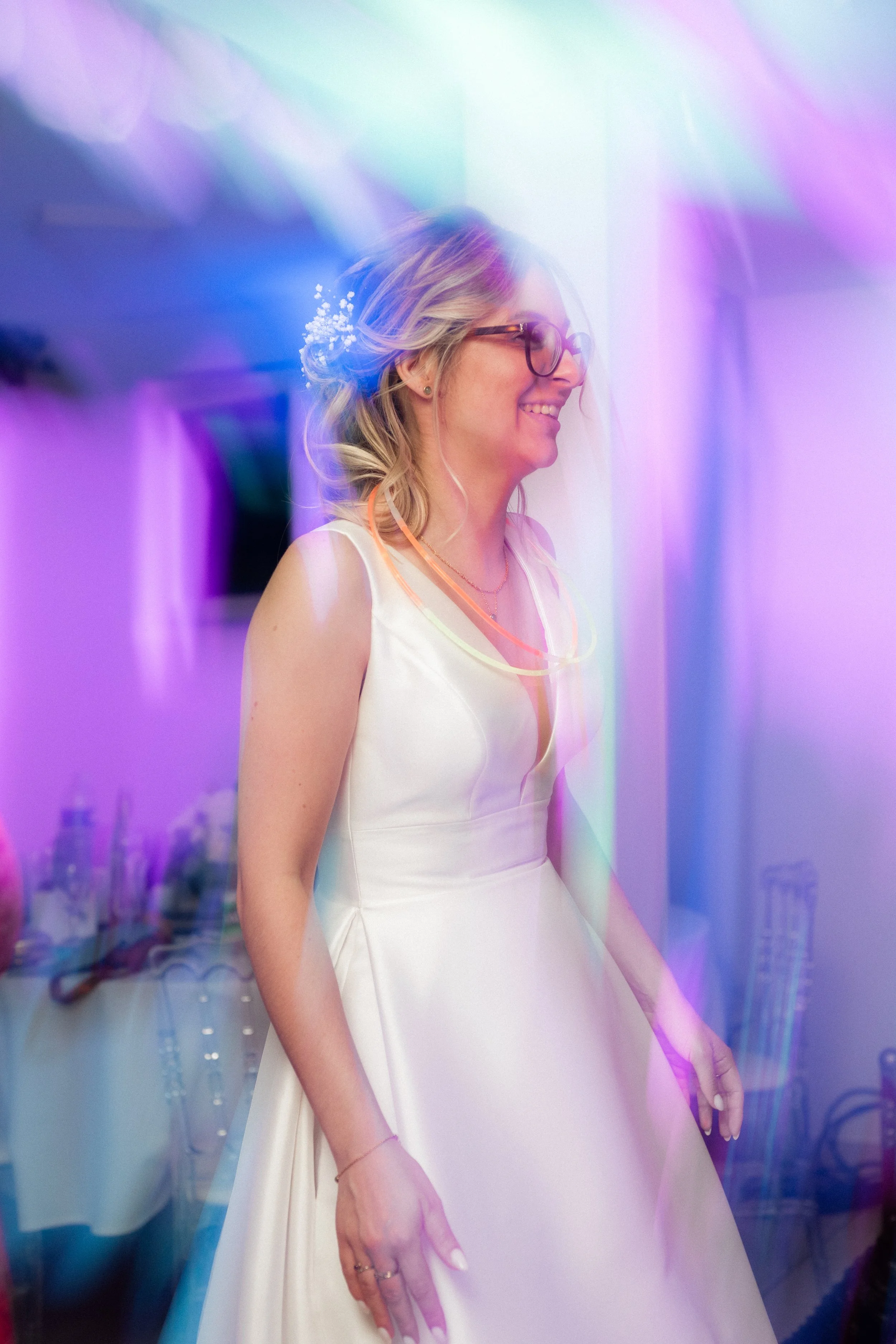 Femme souriante portant une robe de mariée blanche avec une décoration de cheveux et des lunettes, entourée de lumières colorées dans une ambiance festive.