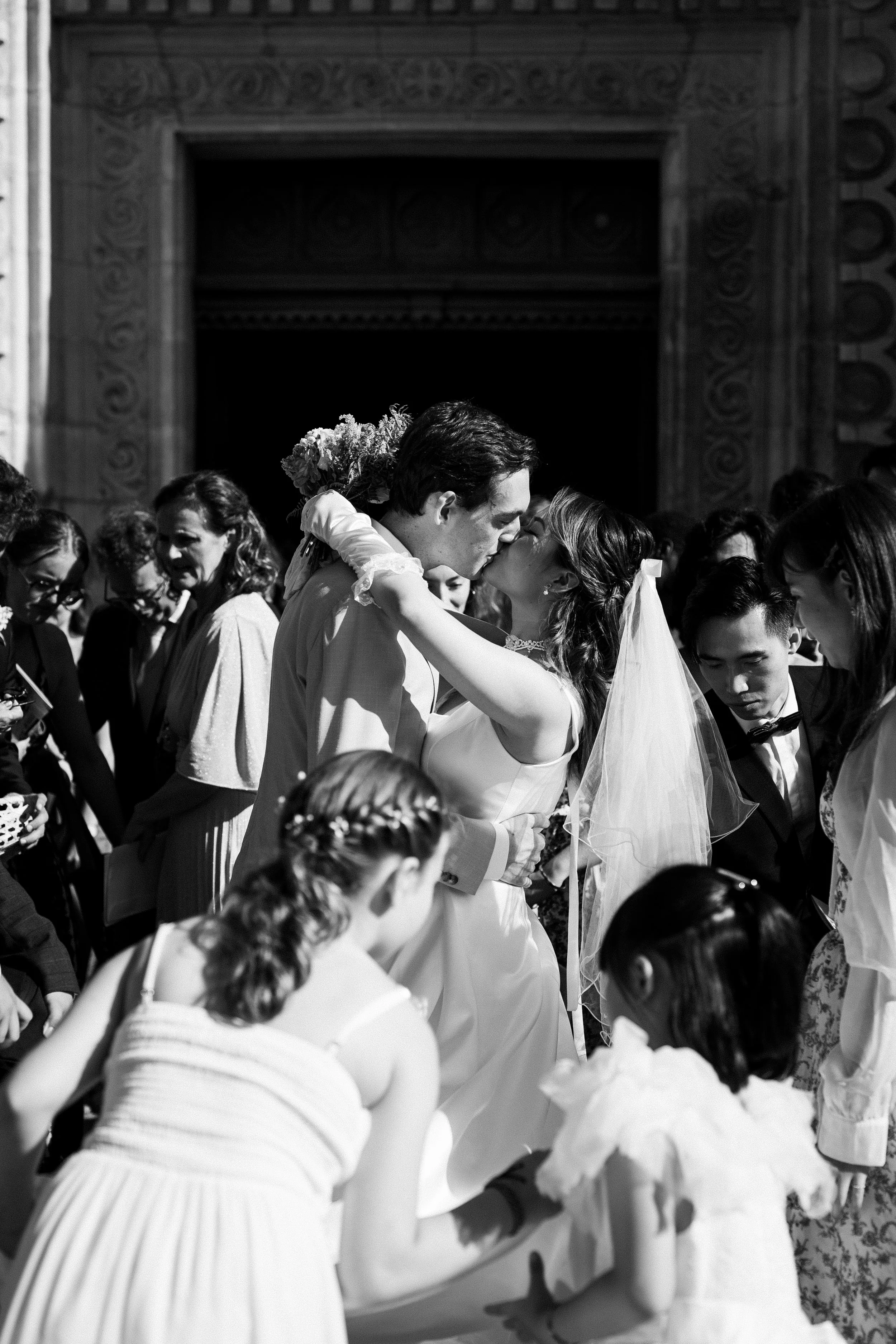 Un couple de mariés s'embrasse lors de leur mariage, entourés d'invités, dans une ambiance festive en noir et blanc.