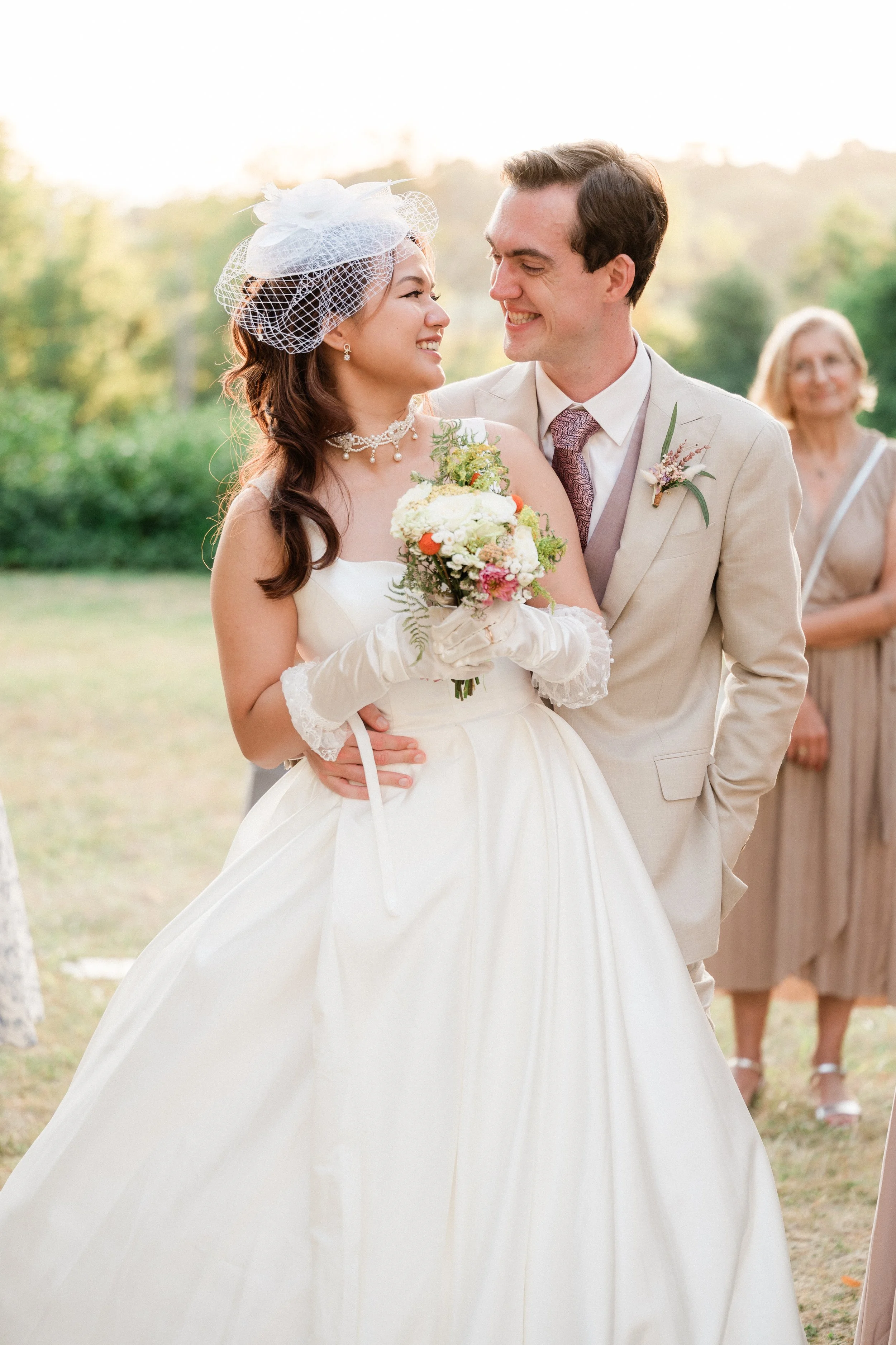 Un couple de mariés souriant lors de leur mariage en extérieur, avec une femme en arrière-plan.