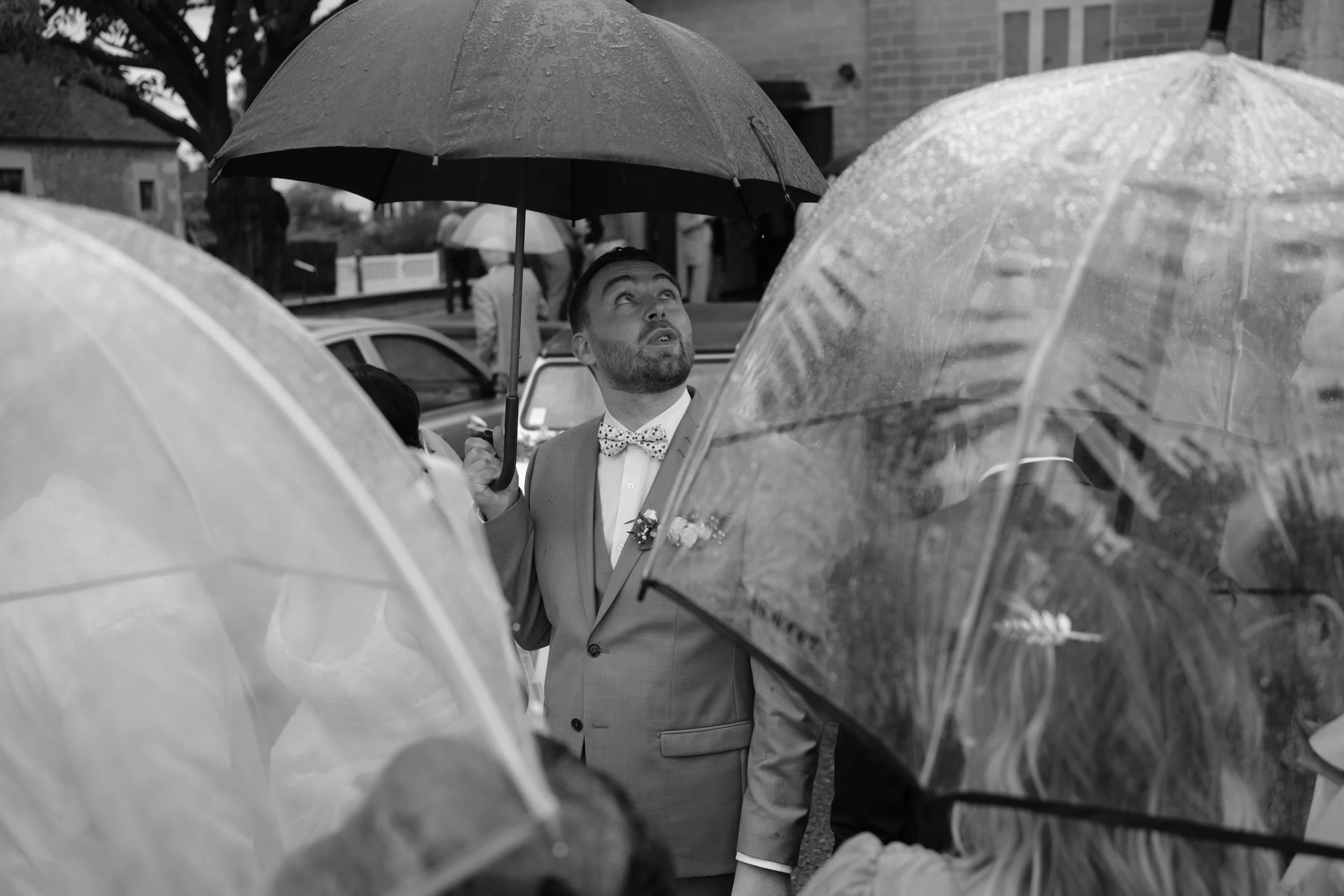 Un homme en costume avec un nœud papillon à pois, regardant vers le haut, entouré de personnes tenant des parapluies par temps de pluie.