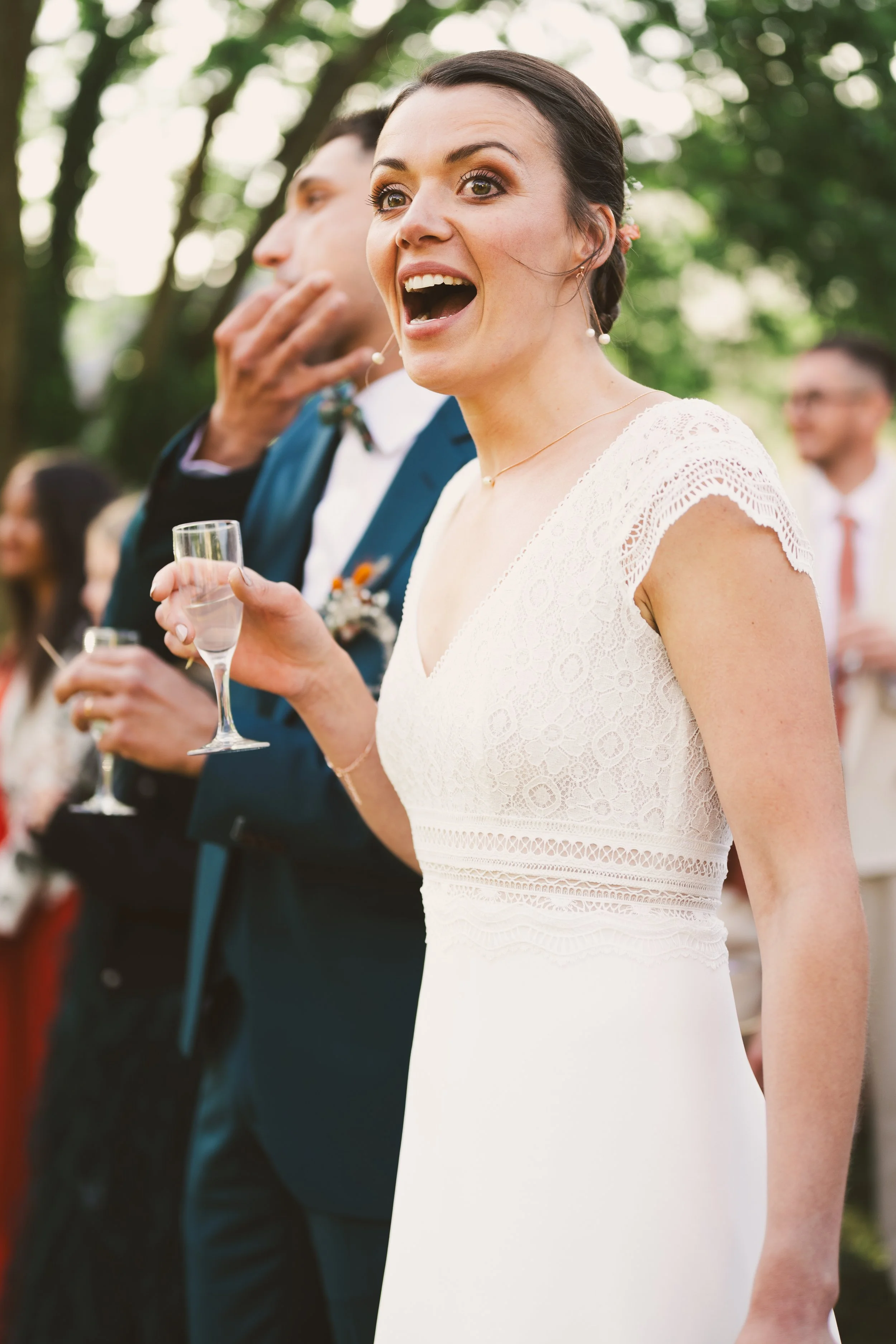 Une femme souriante en robe blanche de mariage tenant une flute de champagne lors d'une célébration en plein air, entourée de personnes en costume.