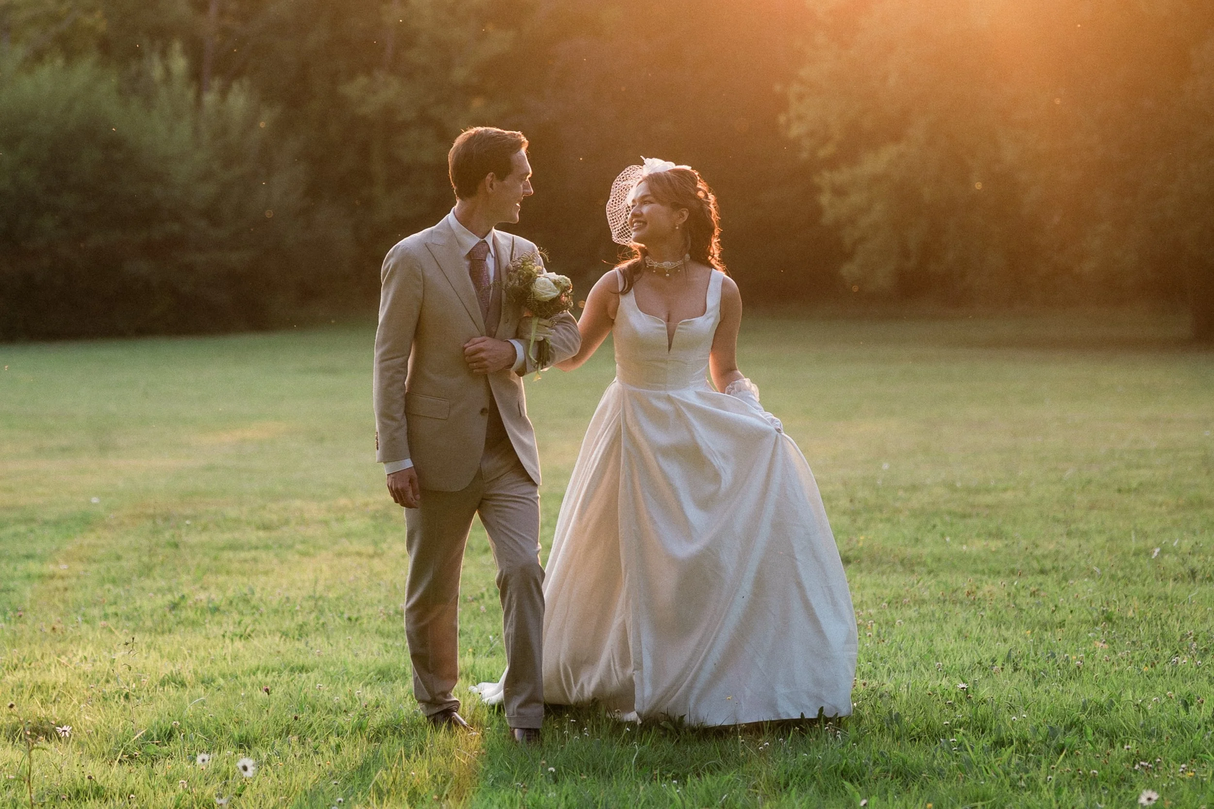 Un couple de mariés marchant dans un champ lors d'un coucher de soleil, la femme portant une robe de mariée blanche et le homme un costume beige. Ils se regardent en souriant.