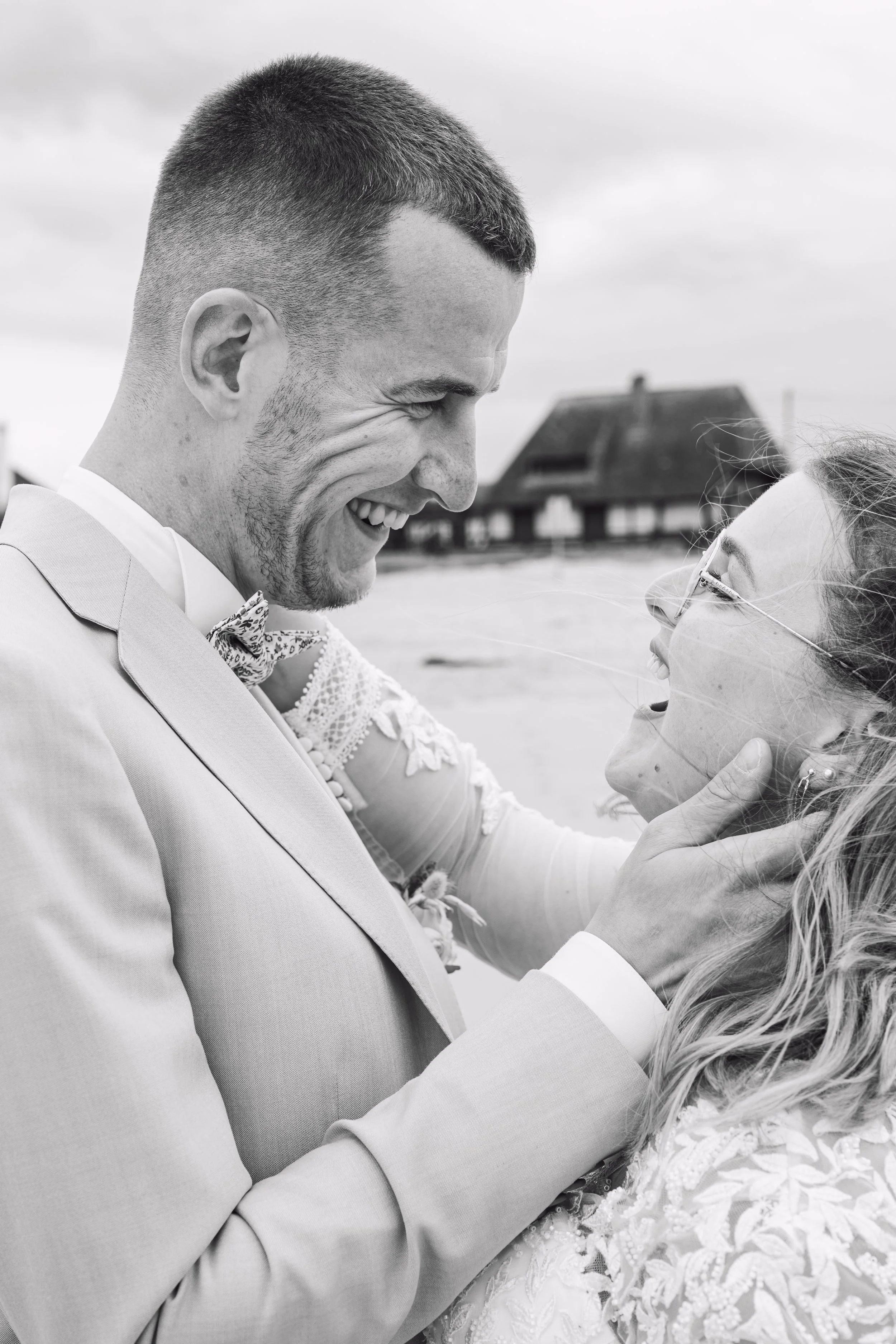 Un couple de mariage, souriant, en gros plan, en noir et blanc, avec une maison en arrière-plan.