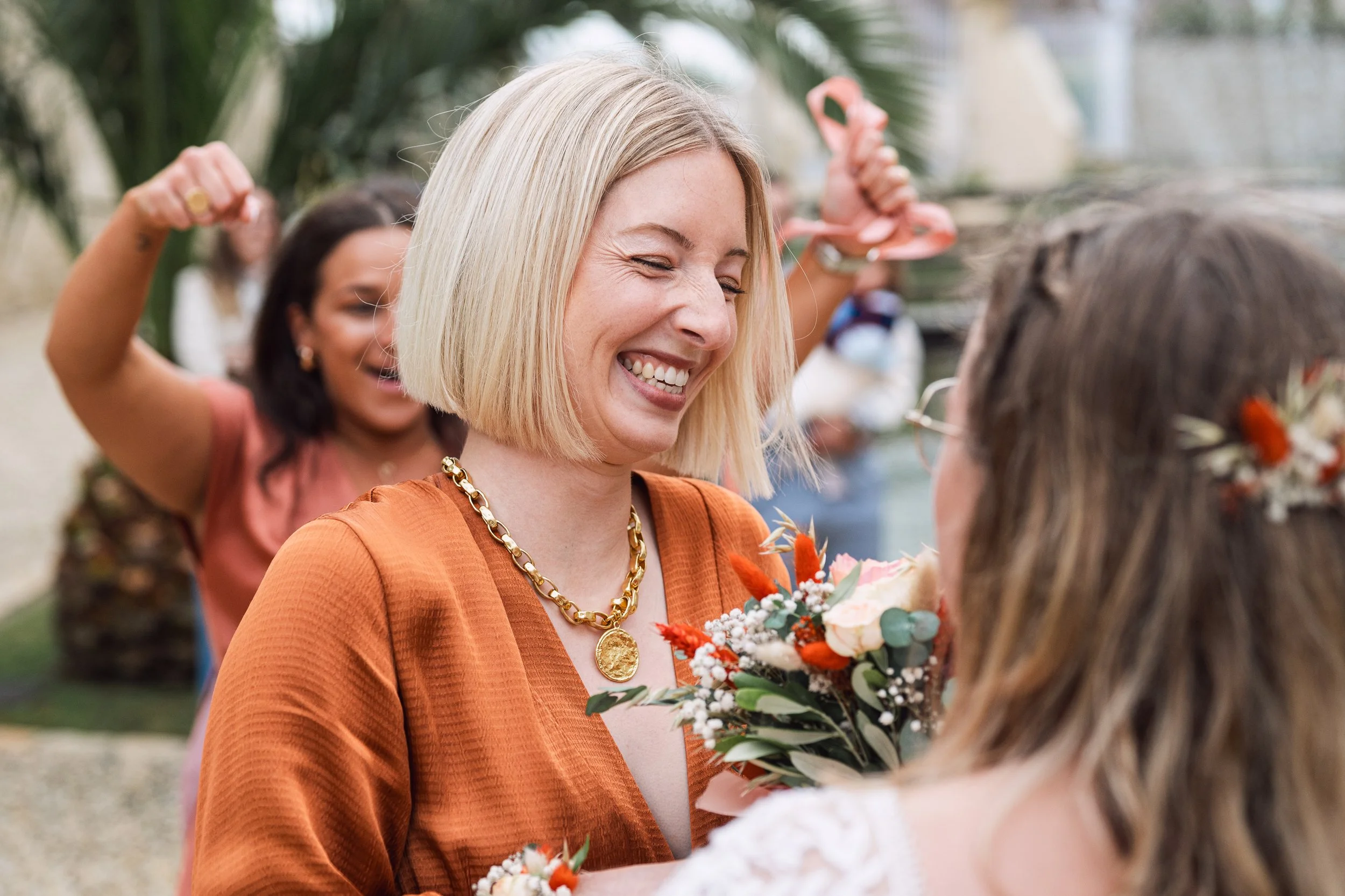 Femme souriante avec un collier en or, tenant un bouquet de fleurs, échangeant un regard avec une autre femme lors d'une cérémonie, avec un groupe de personnes en arrière-plan dans un environnement extérieur.