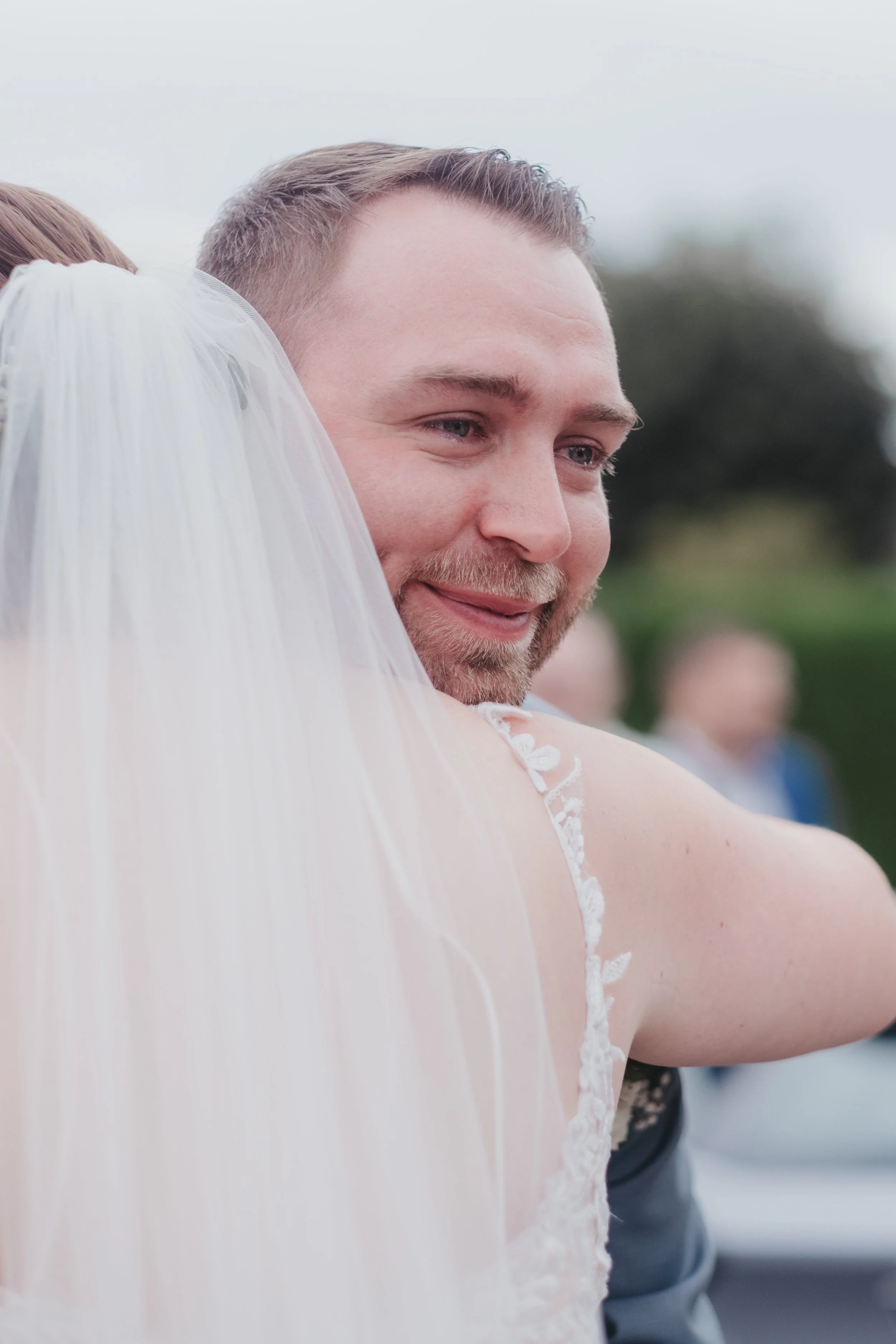 Un homme et une femme en costume de mariage, le sourire, lors d'une cérémonie extérieure.
