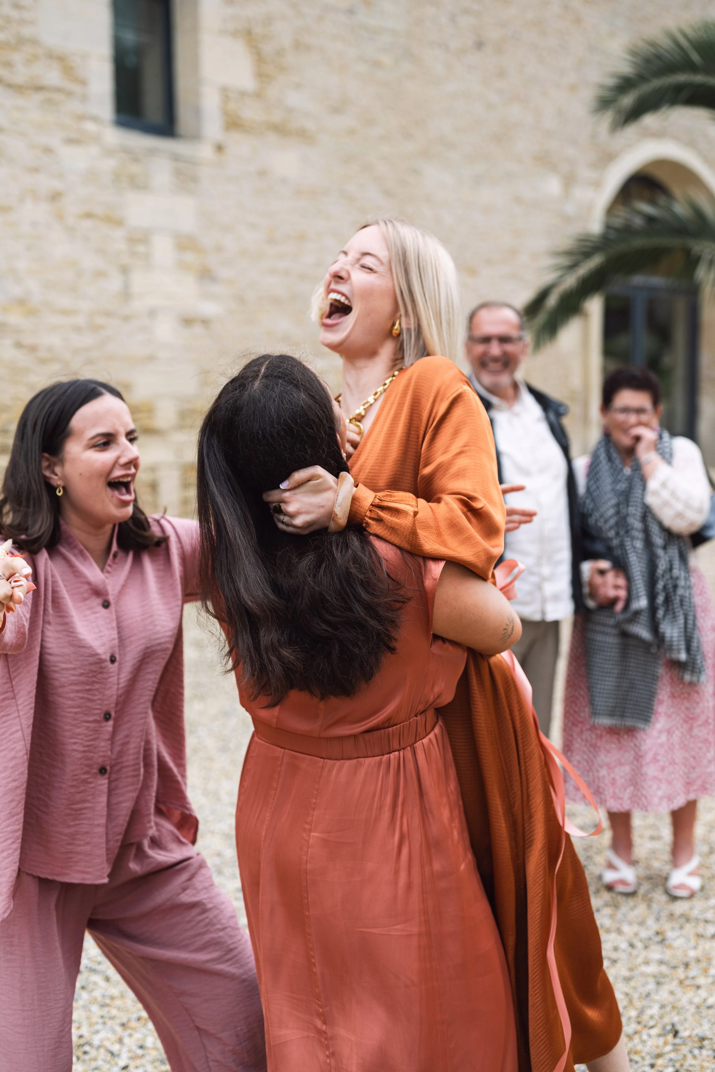 Groupe de personnes souriantes et heureuses lors d'une célébration ou d'une fête à l'extérieur, avec une femme blonde en haut d'une autre femme, tous habillés en vêtements aux tons chauds.