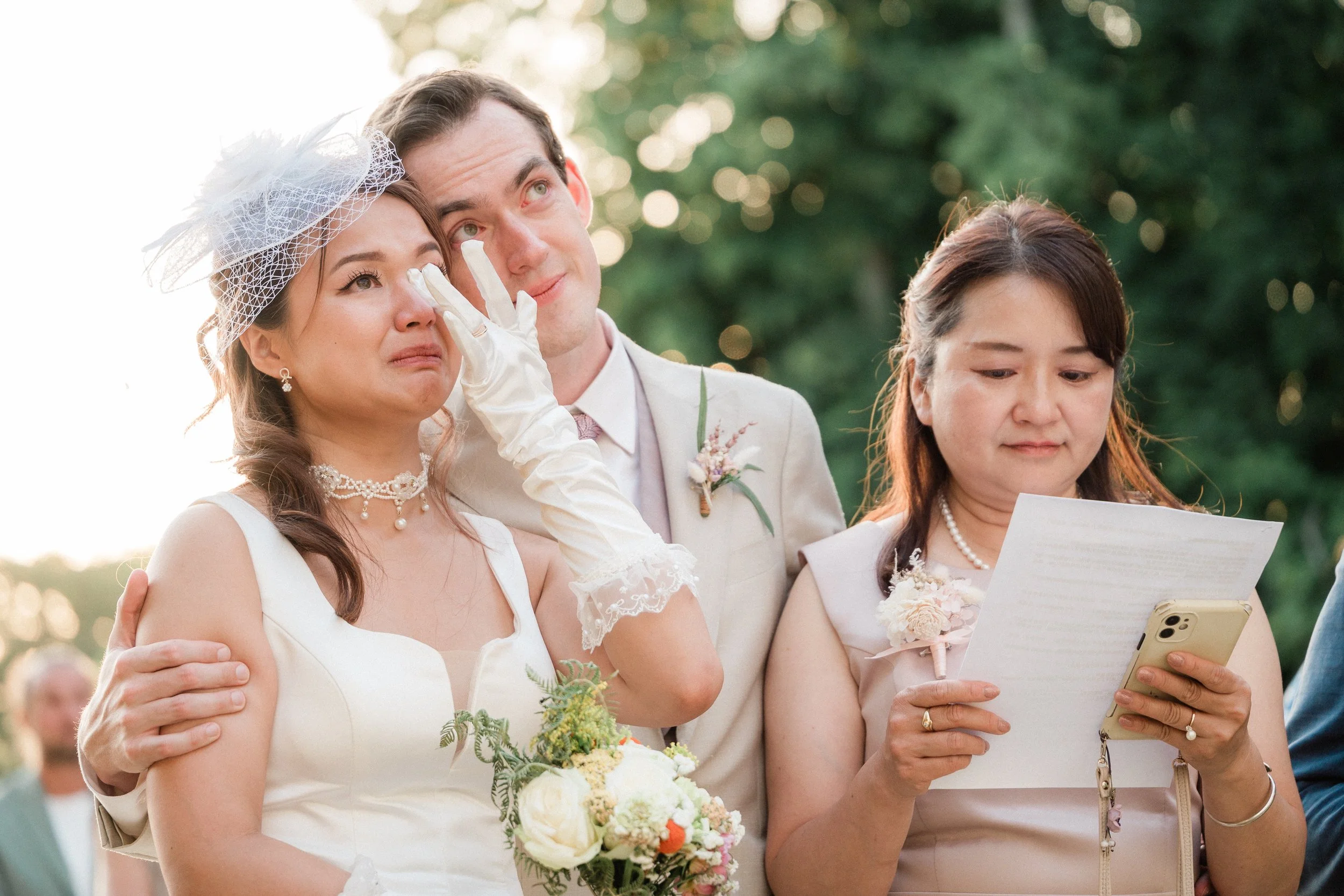 Une jeune mariée est en larmes en pleurant lors d'une cérémonie de mariage. Elle est habillée en blanc avec un collier et un chapeau, et elle pleure en s'essuyant les yeux. À côté d'elle, un homme en costume blanc regarde en larmes, et une femme à cô
