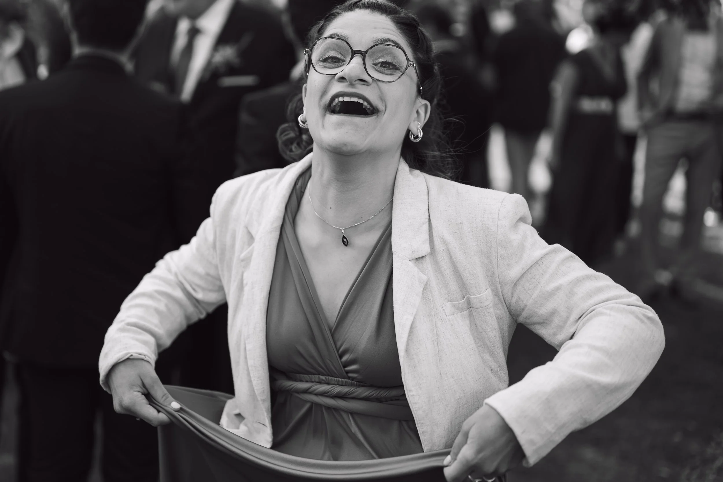 Une femme souriante portant des lunettes, un collier et une veste claire, en train de rire ou de s'amuser, avec des personnes en arrière-plan.