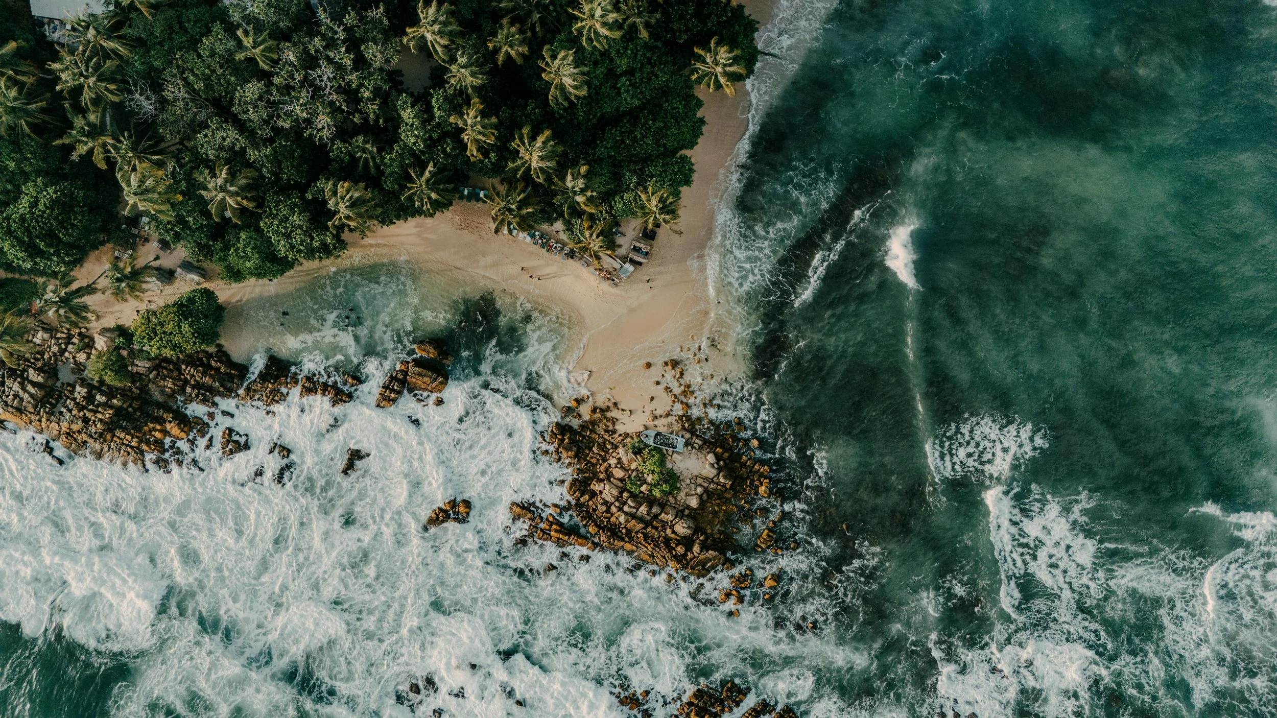 Aerial view of a tropical beach with dense palm trees and lush greenery, sandy shoreline, rocks, and ocean waves.