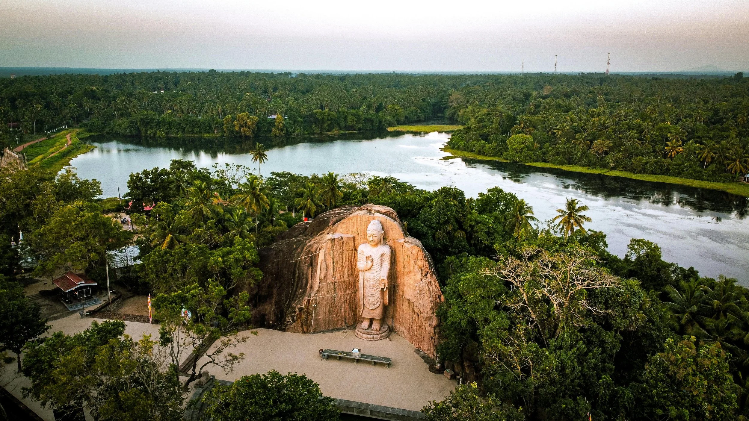 A large carved Buddha statue situated at the base of a rock formation, surrounded by lush greenery and a river in the background.