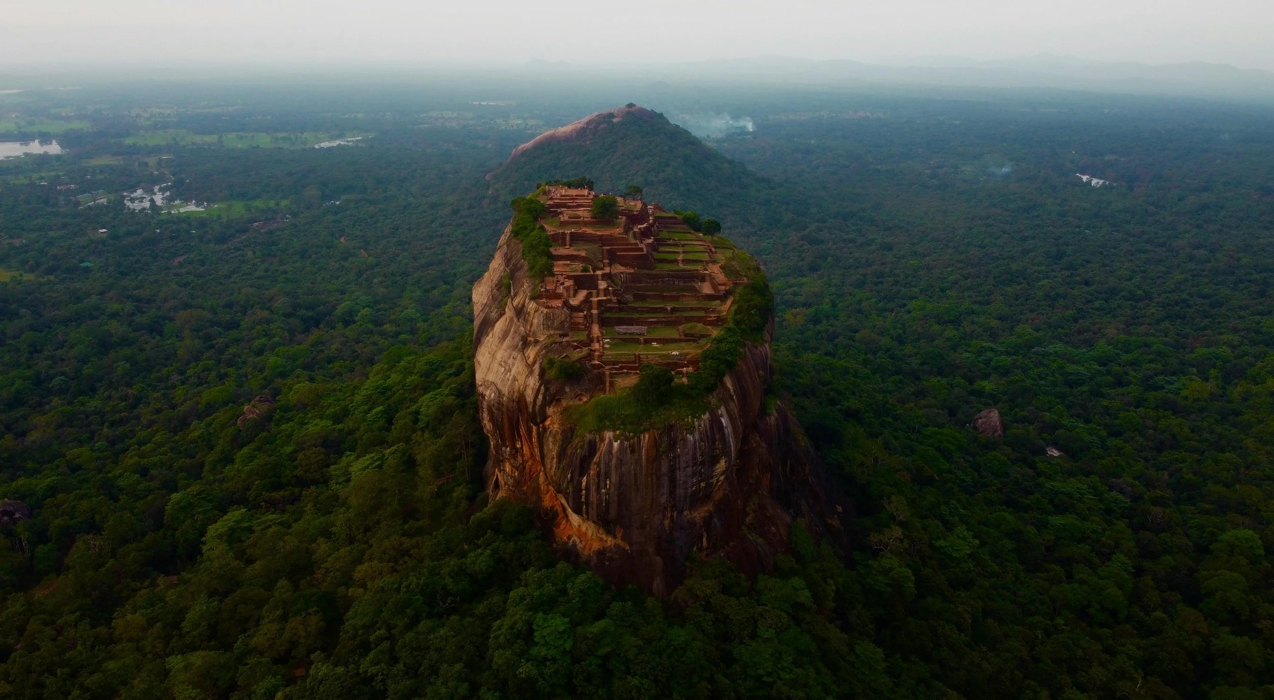 A large rock formation with ancient terraced buildings on top, surrounded by dense green forest, with rolling hills and cloudy sky in the background.