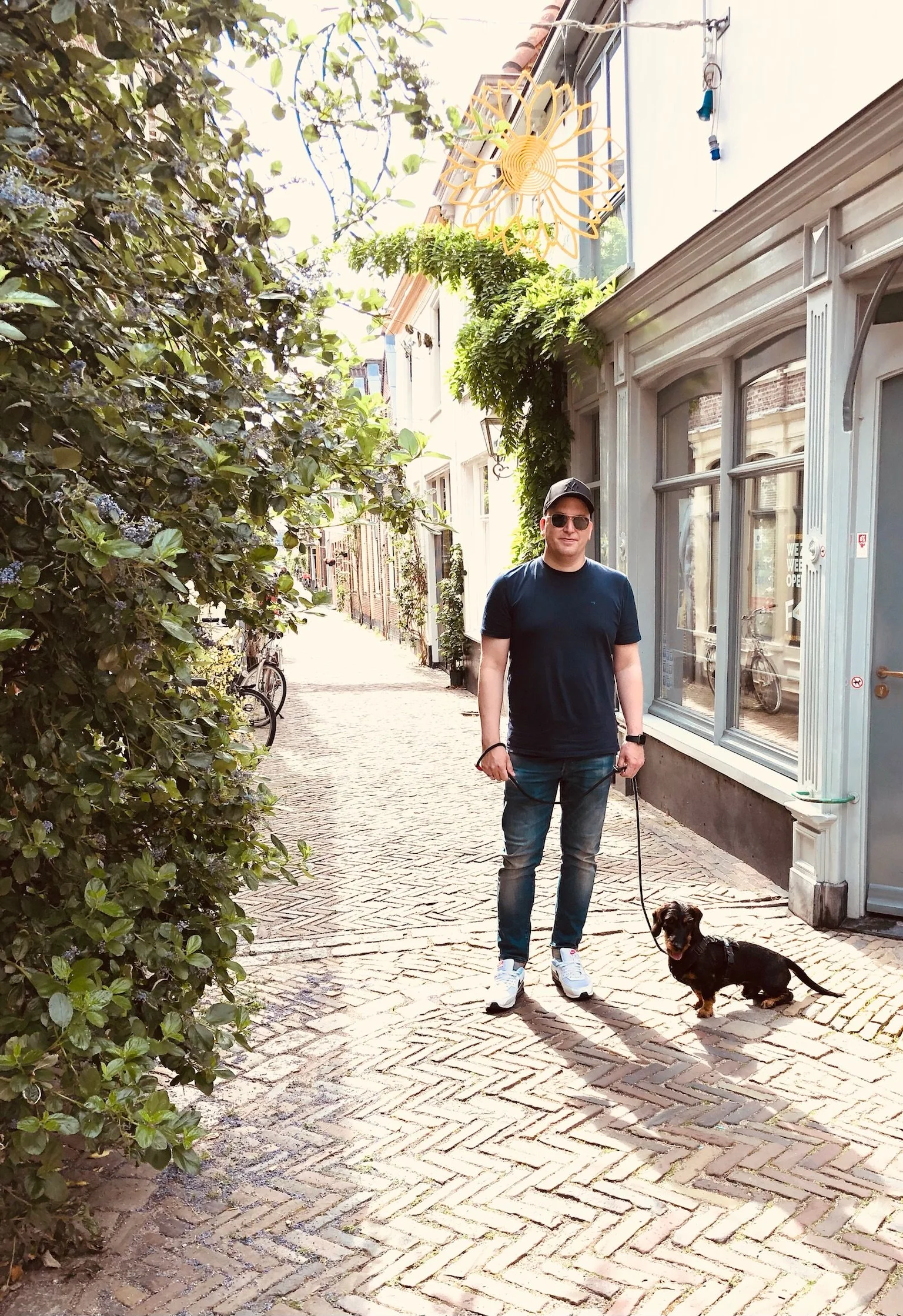 Ein Mann auf einer Kopfsteinpflasterstraße mit einem kleinen Hund an der Leine, umgeben von einem urbanen Wohnviertel mit Gebäuden und Büschen.