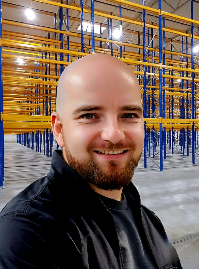 Ein Mann mit einem Bart und Glatzkopf vor einem Lagerregal im Lagerhaus.