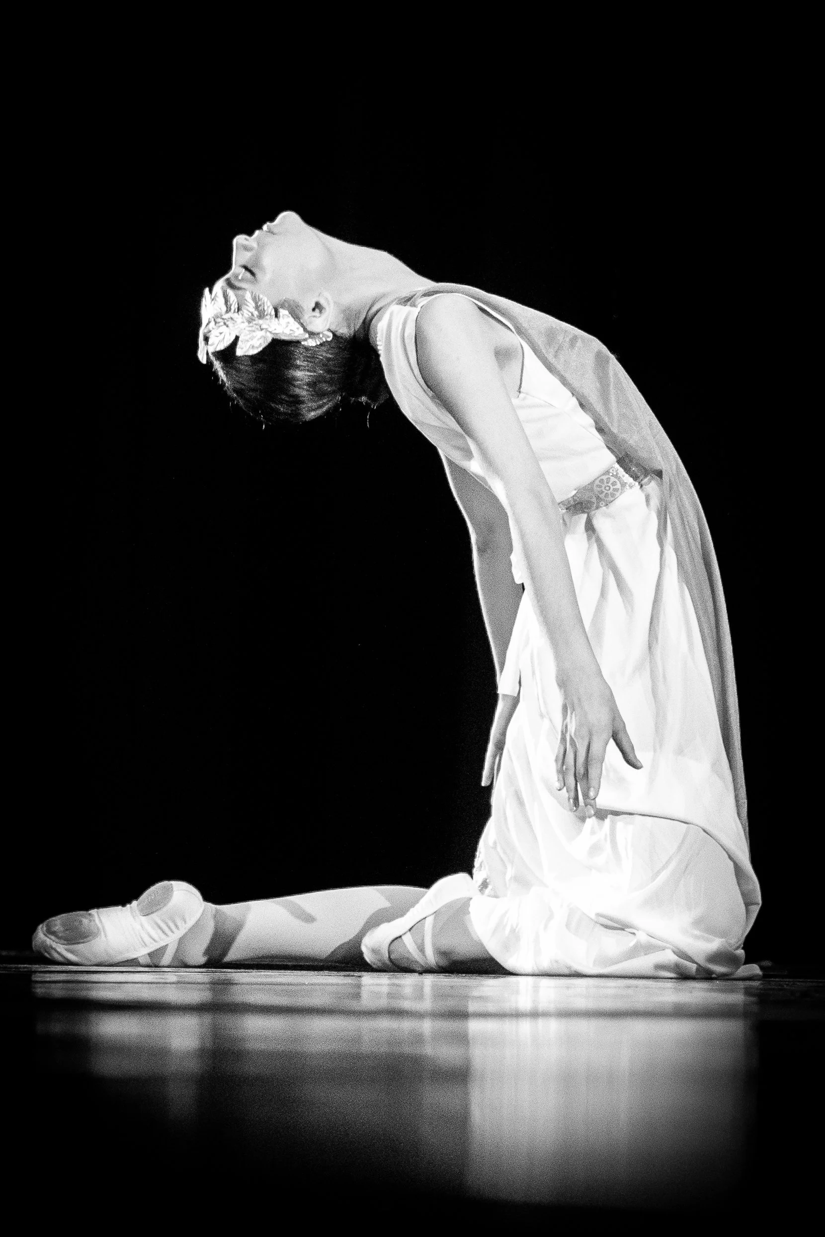 Une ballerine en costume blanc, agenouillée sur scène avec le dos arqué et la tête inclinée en arrière, en noir et blanc.