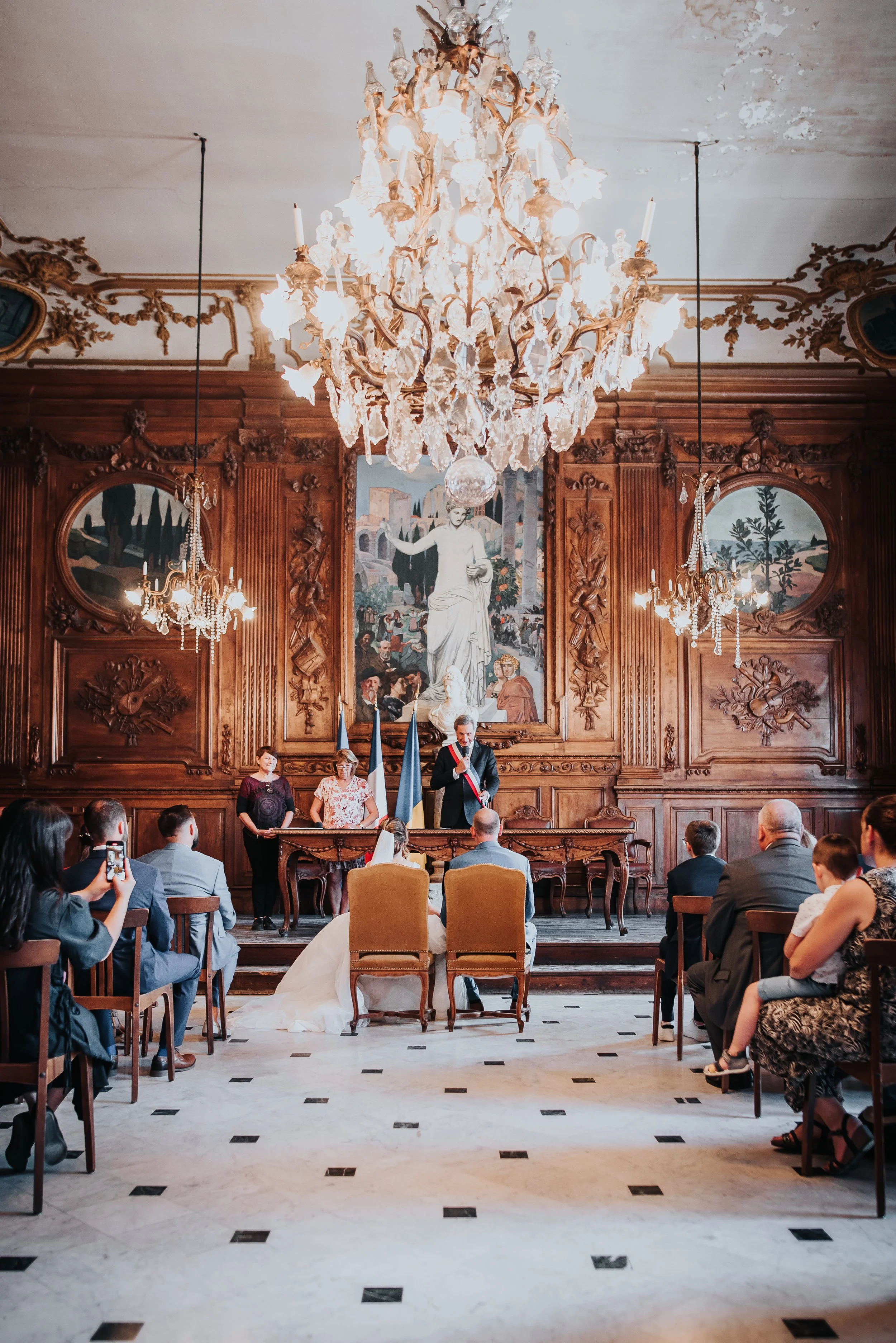 Cérémonie de mariage dans une salle ornée de bois sculpté, avec un chandelier chandelier en cristal au plafond, un tableau mural, et des personnes assises, en train de prendre des photos.