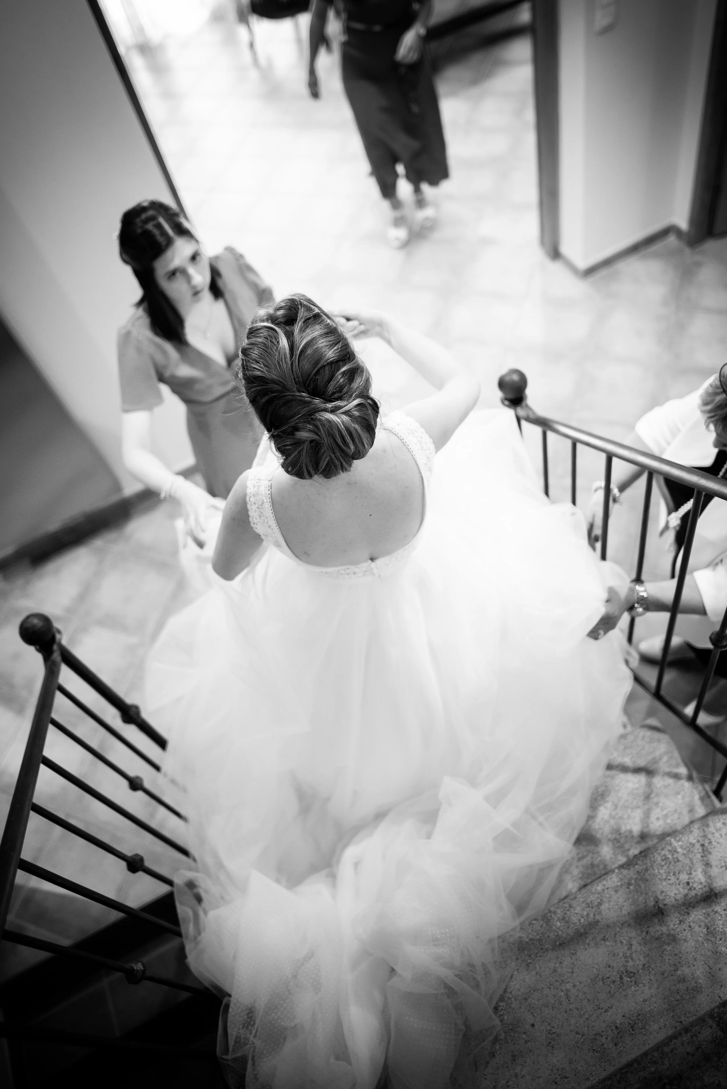Une femme en robe de mariée descend un escalier en fer forgé, vue depuis le dessus, avec deux autres femmes qui l'aident ou discutent avec elle.