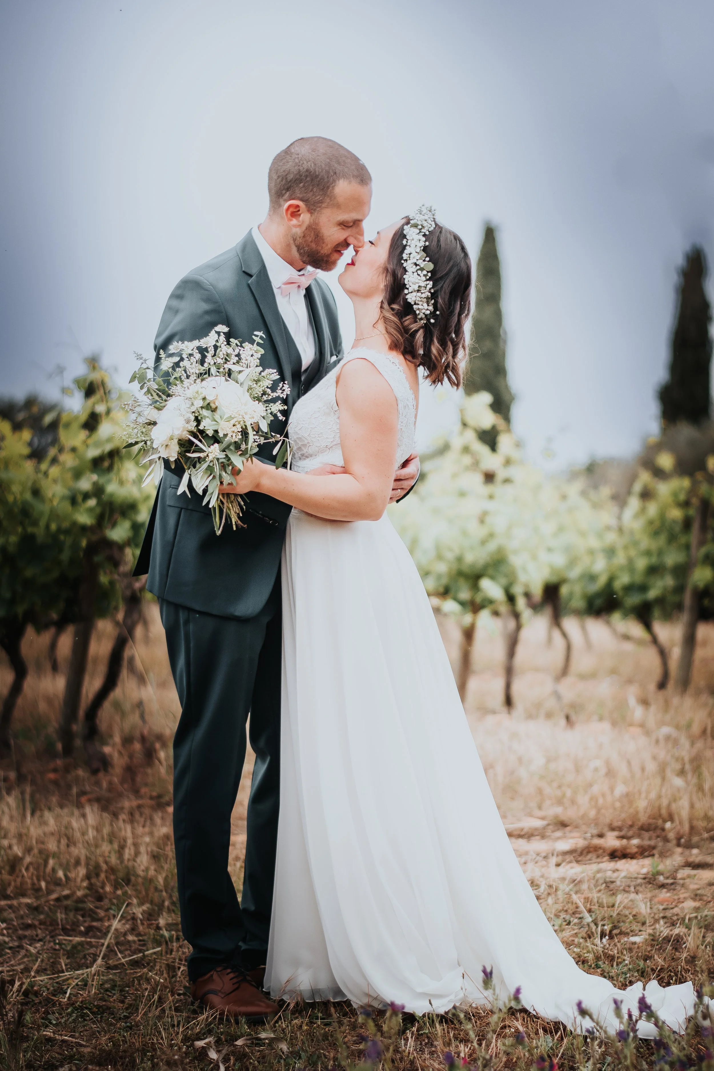 Un couple de mariés s'embrassant dans un vignoble, l'homme en costume gris et la femme en robe de mariée blanche avec une couronne de fleurs, par temps nuageux.