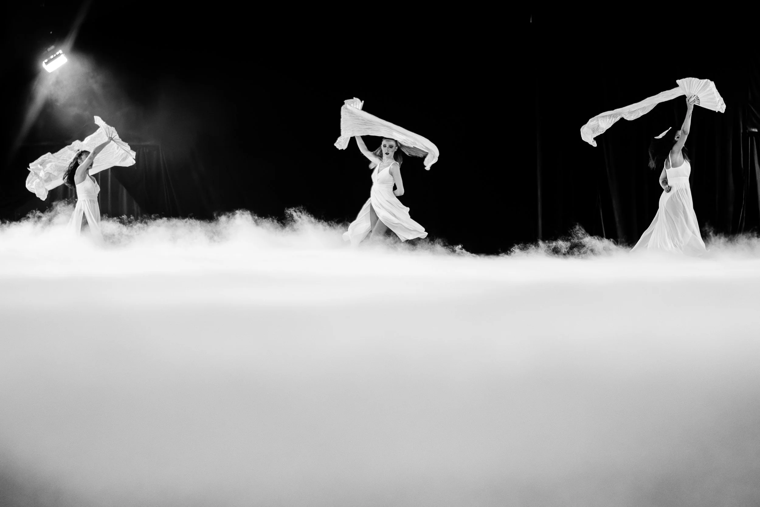 Trois femmes en robes longues blanches dansant avec des voiles, dans un espace sombre avec de la brume au sol.