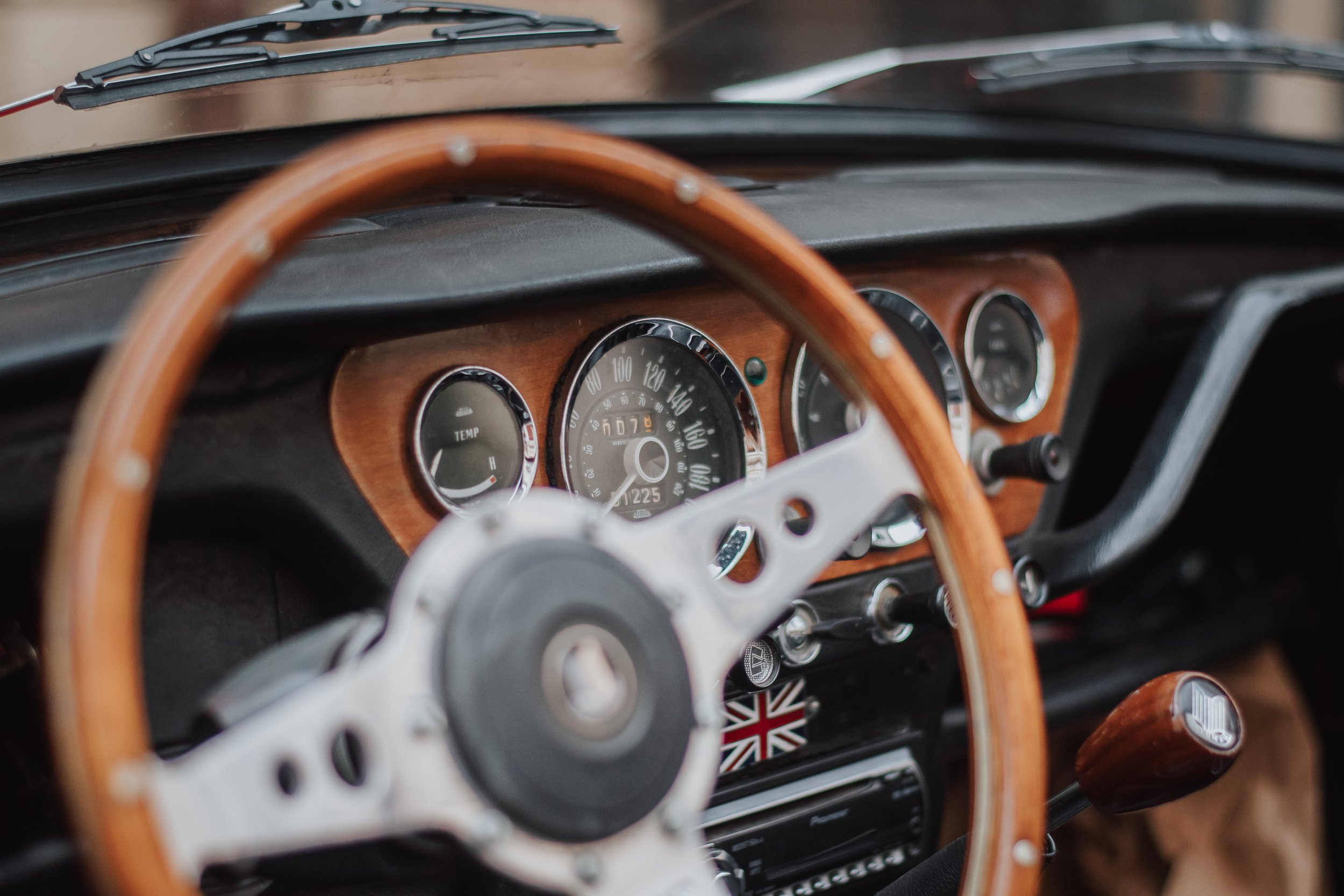 Intérieur d'une voiture vintage avec un tableau de bord en bois, un volant en bois et métal, et un levier de vitesse en bois. La console montre un indicateur de température et un compteur de vitesse.