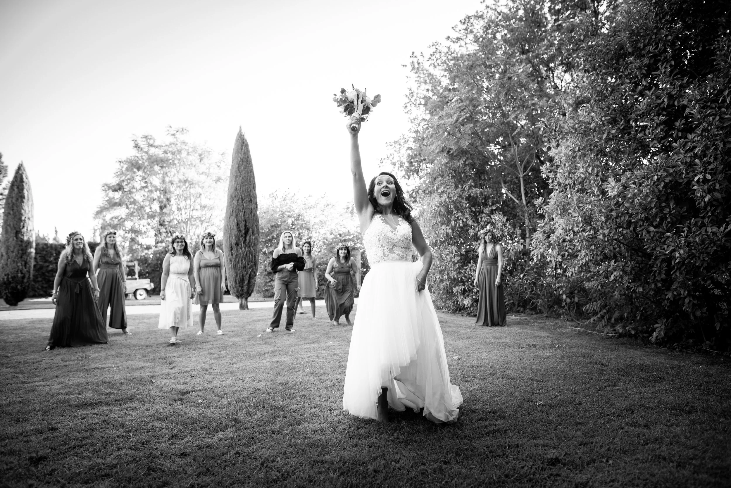 Une mariée en robe blanche lançant un bouquet de fleurs vers ses amis lors d'un mariage en plein air.