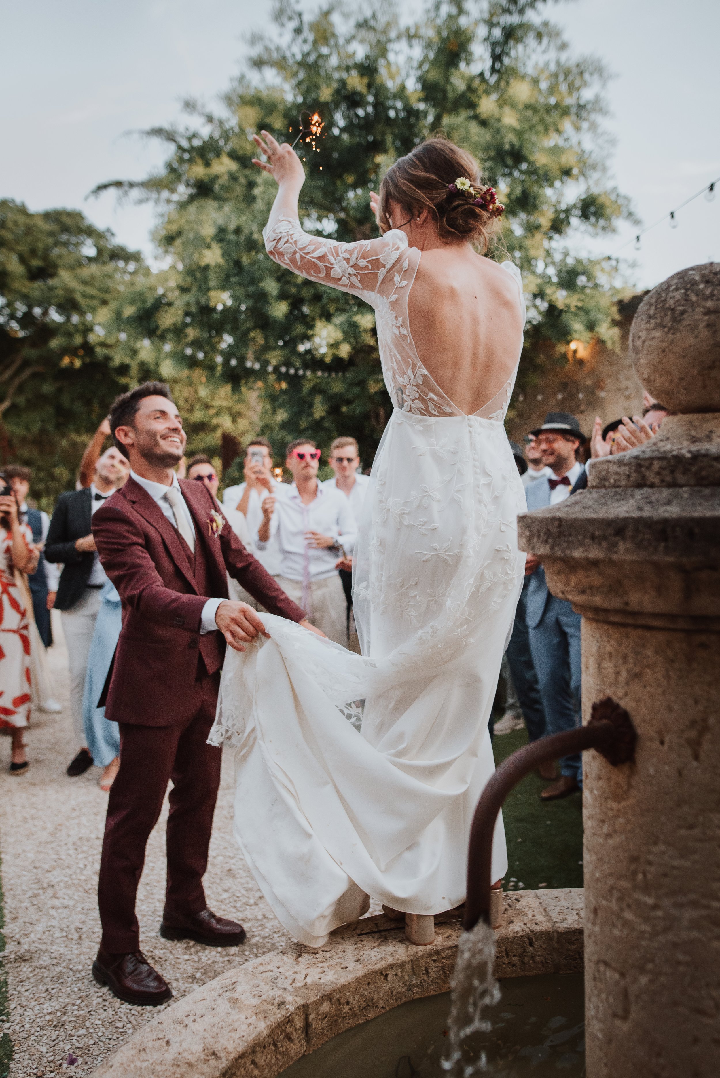 Un couple de mariage, la mariée en robe blanche sirène avec broderies en dentelle, est en l'air devant une fontaine, entourée de convives qui prennent des photos et applaudissent dans un jardin avec des arbres et des guirlandes lumineuses le soir.