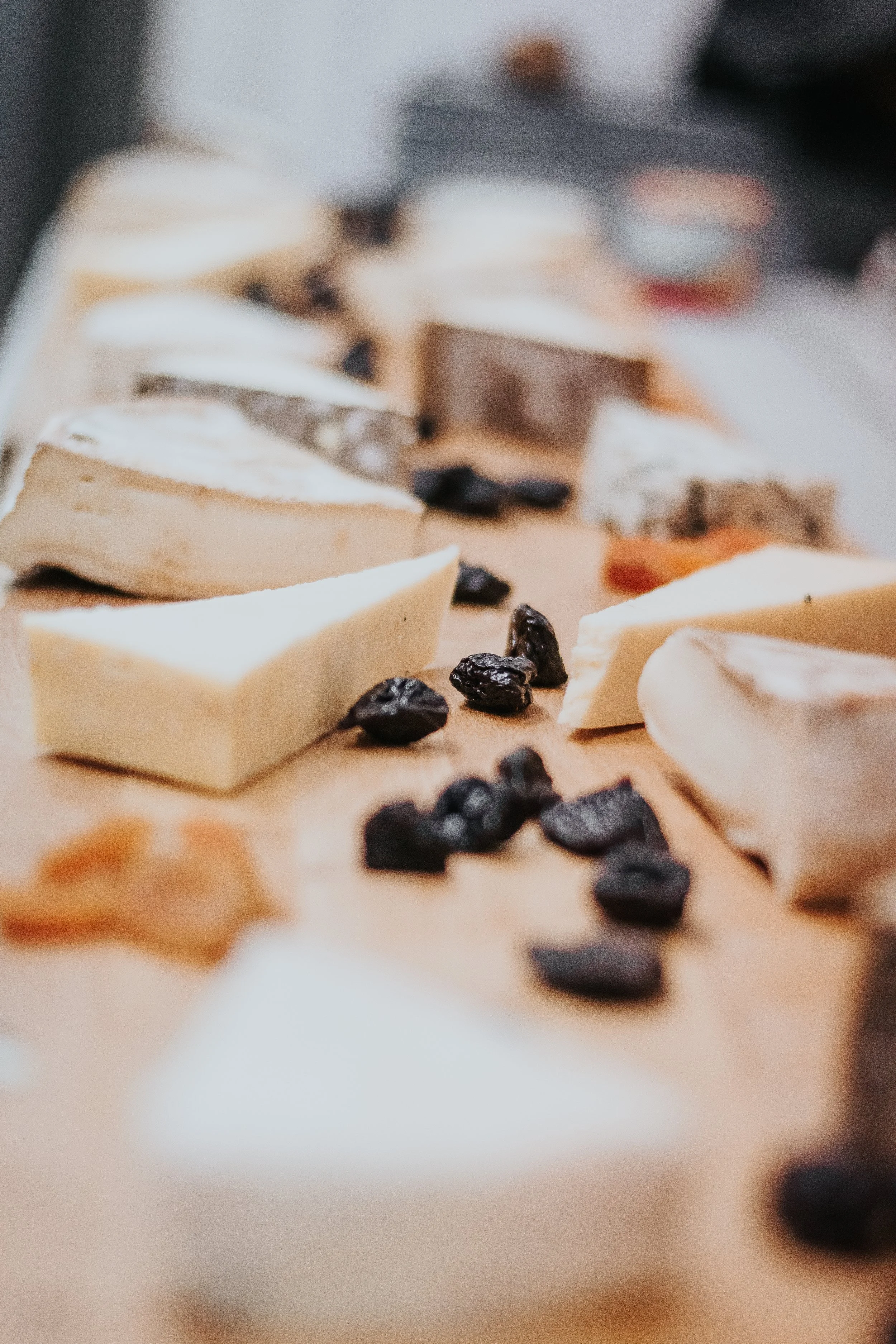 Différents types de fromages placés sur une planche en bois, avec quelques raisins secs autour, image floue en arrière-plan.