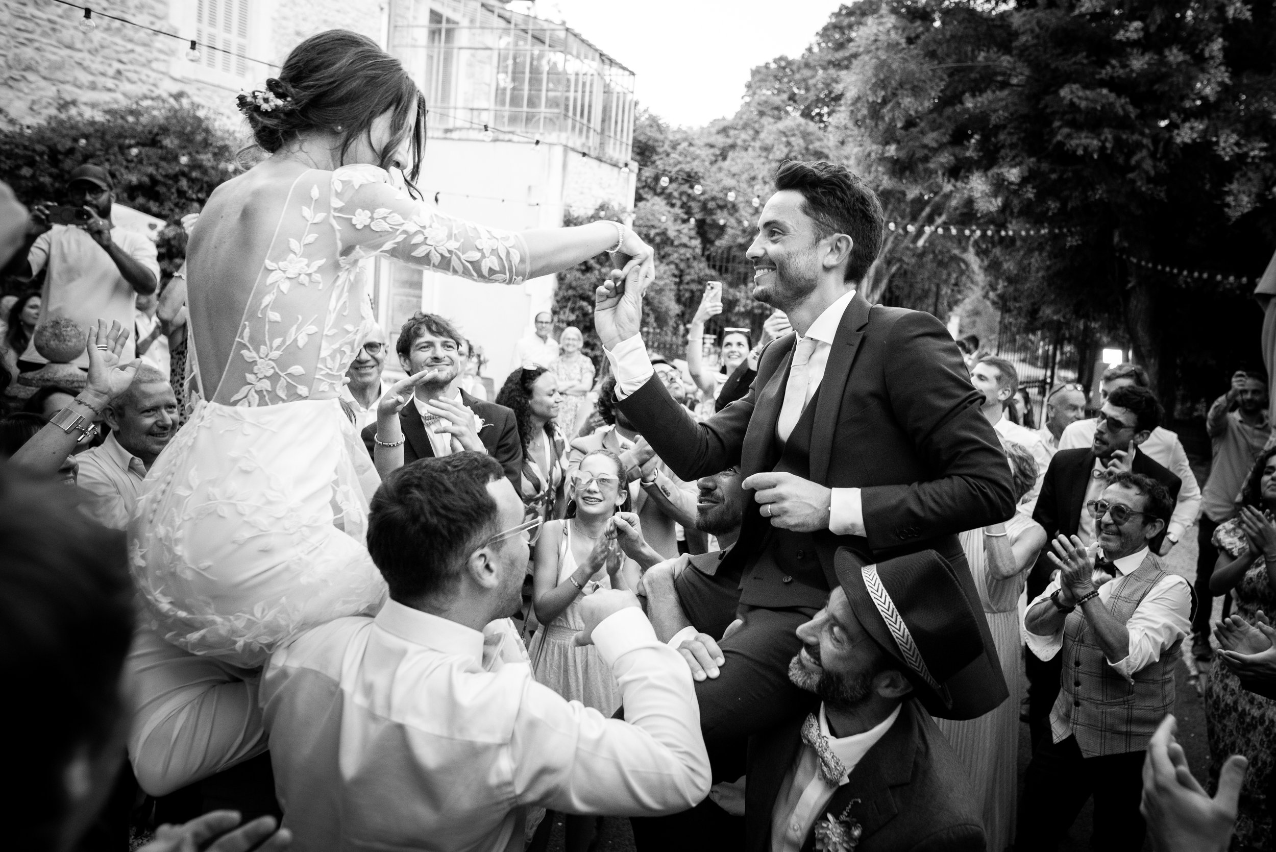 Un couple de mariés lors d'une célébration en plein air, entouré de famille et amis qui applaudissent et prennent des photos, la femme en robe de mariée brodée et l'homme en costume.