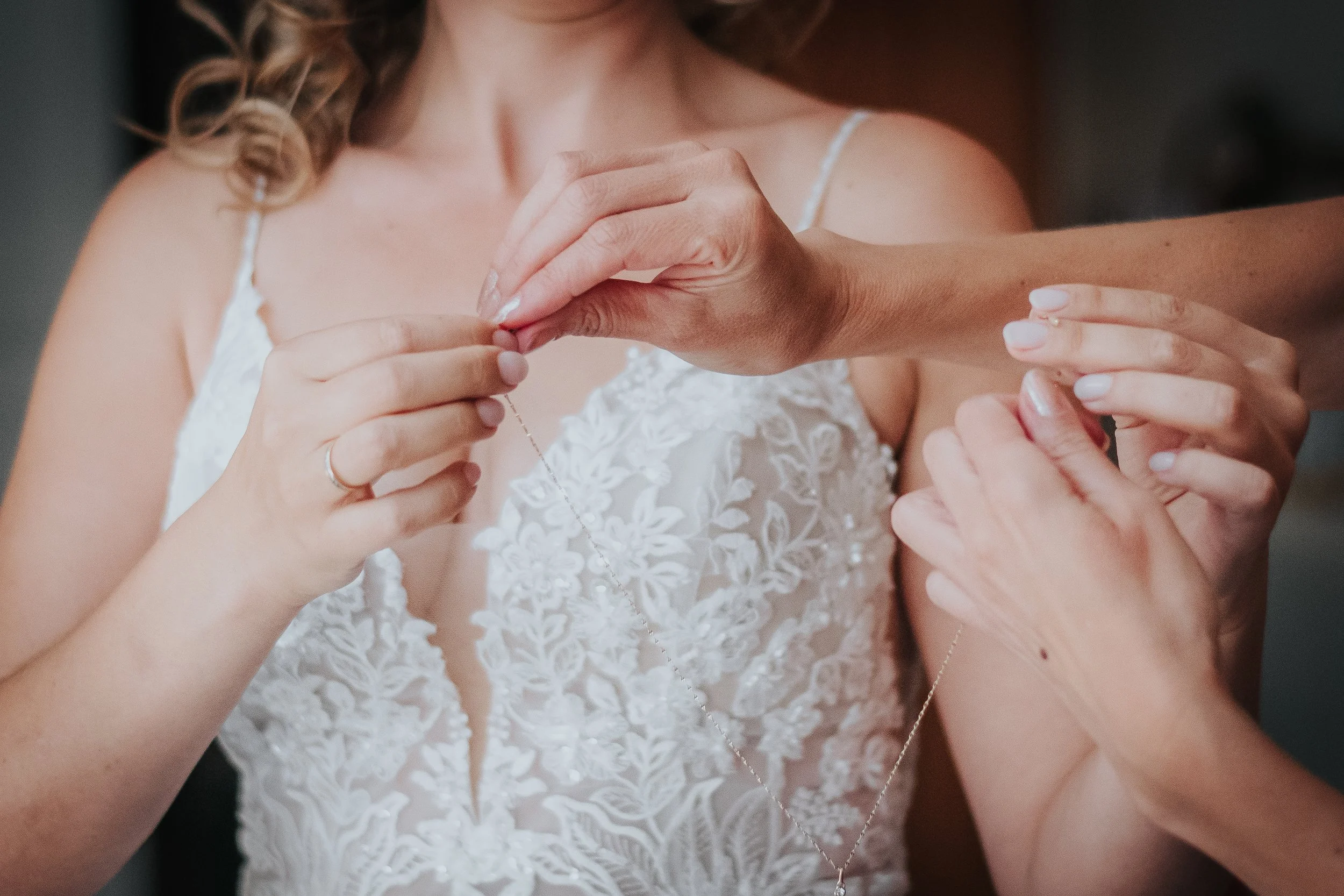 Une femme en robe de mariée est en train de mettre un collier, assistée par d'autres mains.