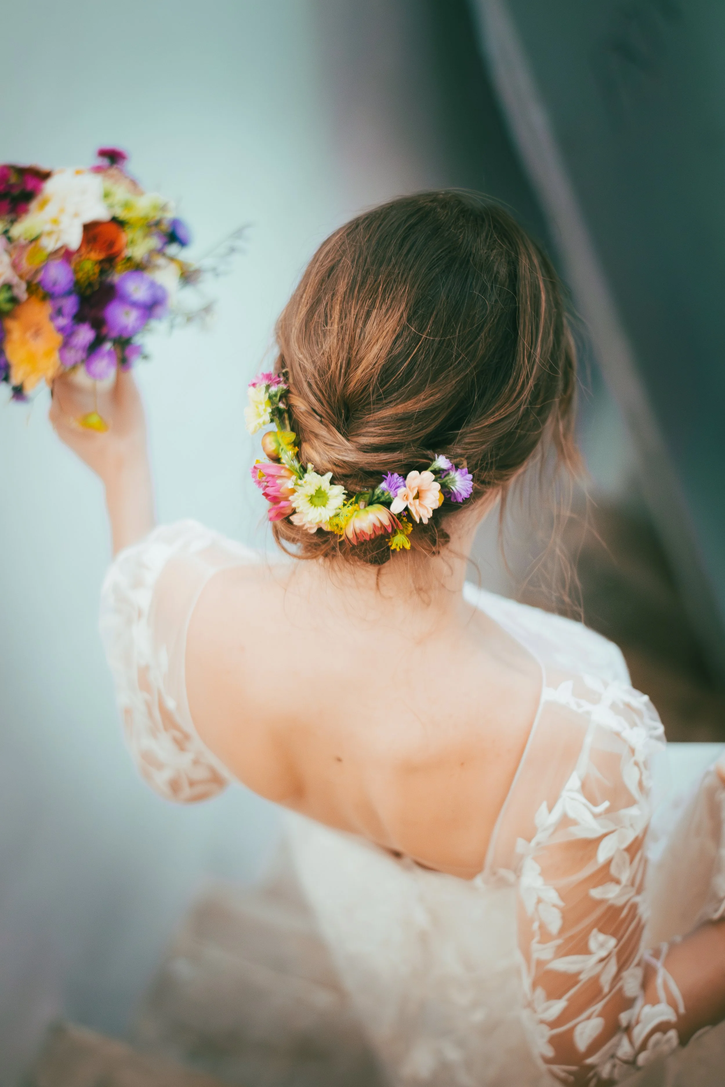 Femme vue de dos portant une couronne de fleurs et une robe en dentelle, tenant un bouquet de fleurs.