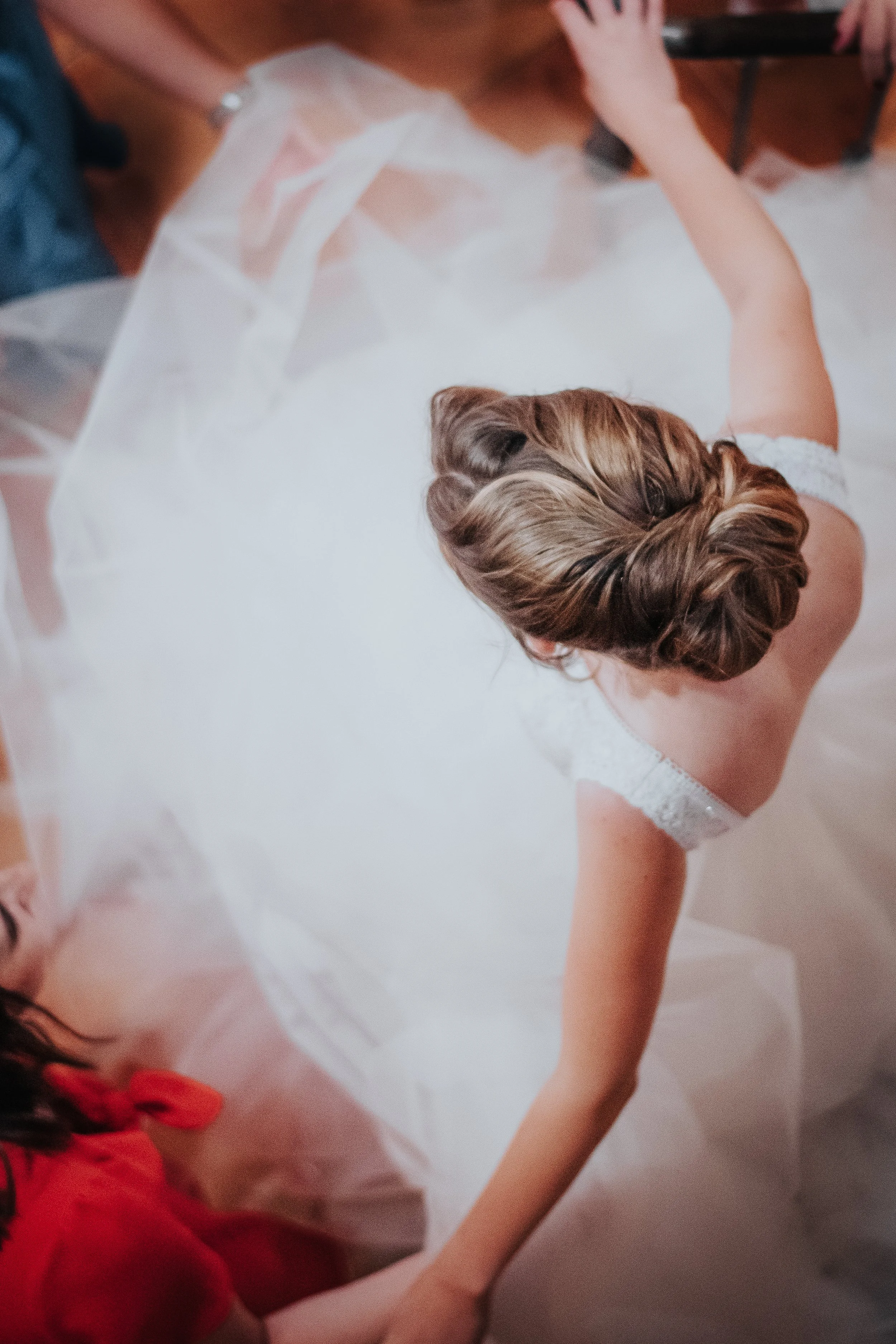 Une femme en robe blanche assise sur le sol, vue du-dessus, avec des cheveux bruns en chignon, entourée de personnes. Il semble s'agir d'une mariée lors d'une préparation ou d'une séance photo.