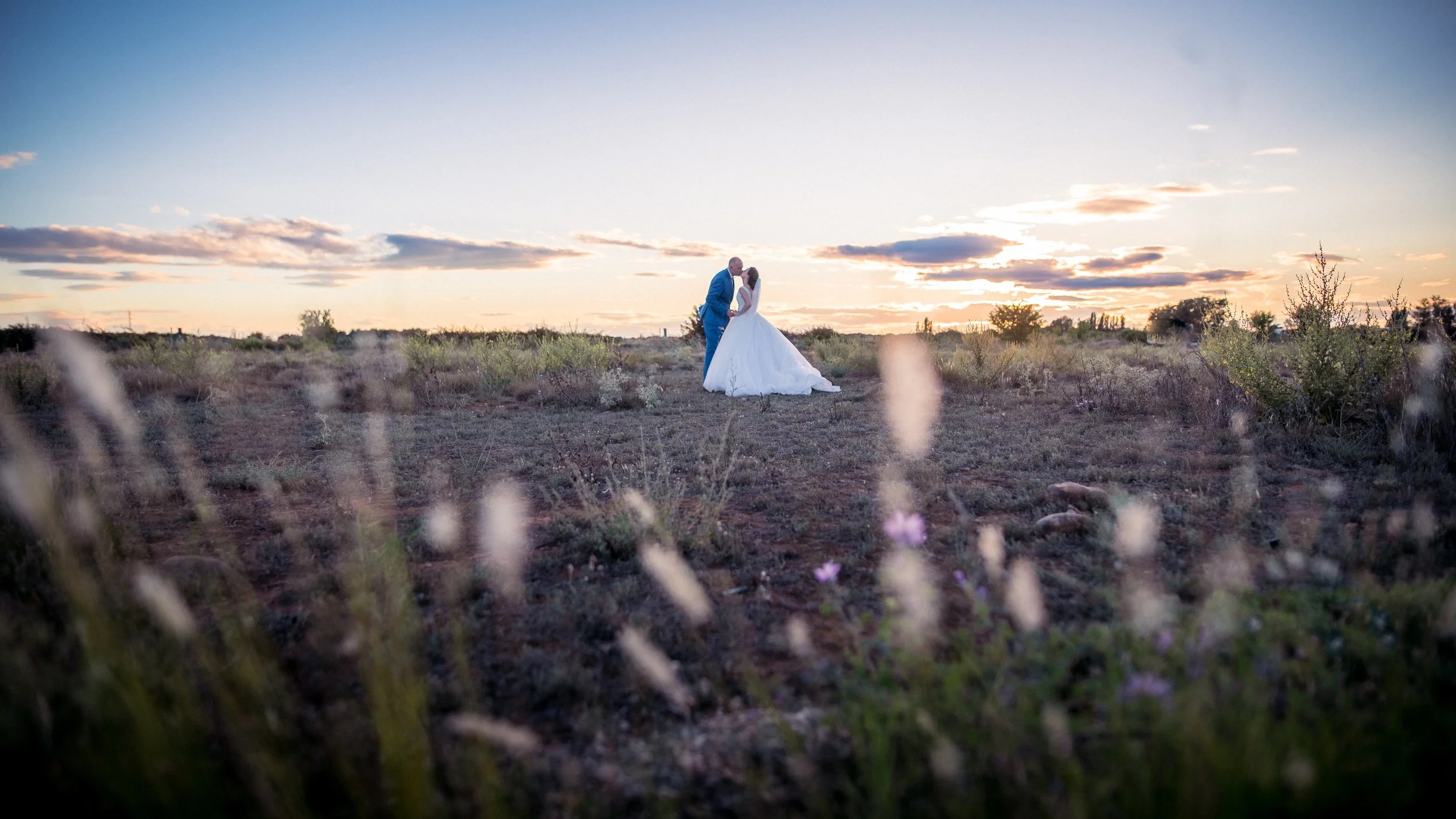 Un couple de mariés danse dans un champ désert au coucher du soleil, avec un ciel coloré en arrière-plan.