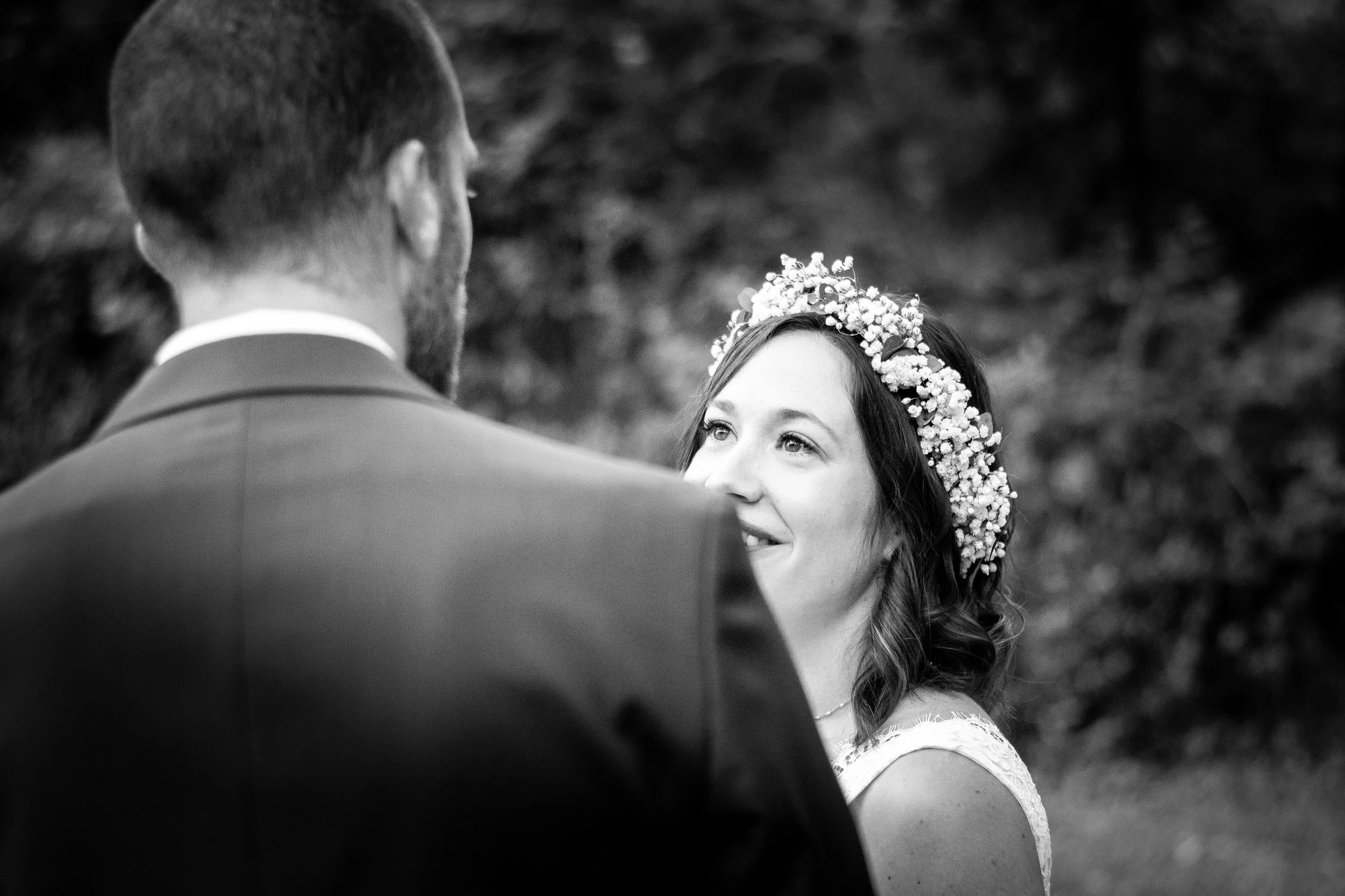 Une femme souriante portant une couronne de fleurs regarde un homme dont le dos est visible. L'image est en noir et blanc, probablement lors d'une cérémonie ou d'un moment intime.photographe mariage Nimes delor alexandre