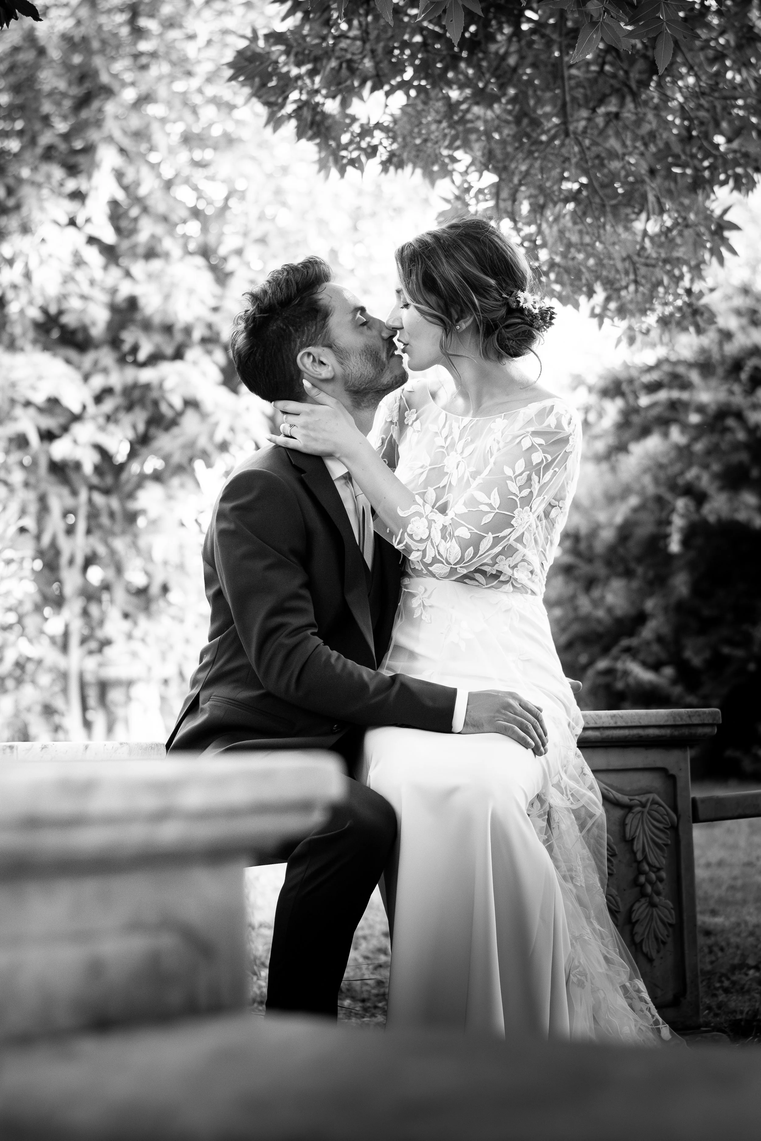 Un couple en tenue de mariage partage un baiser dans un parc, arbres en arrière-plan, photo en noir et blanc.