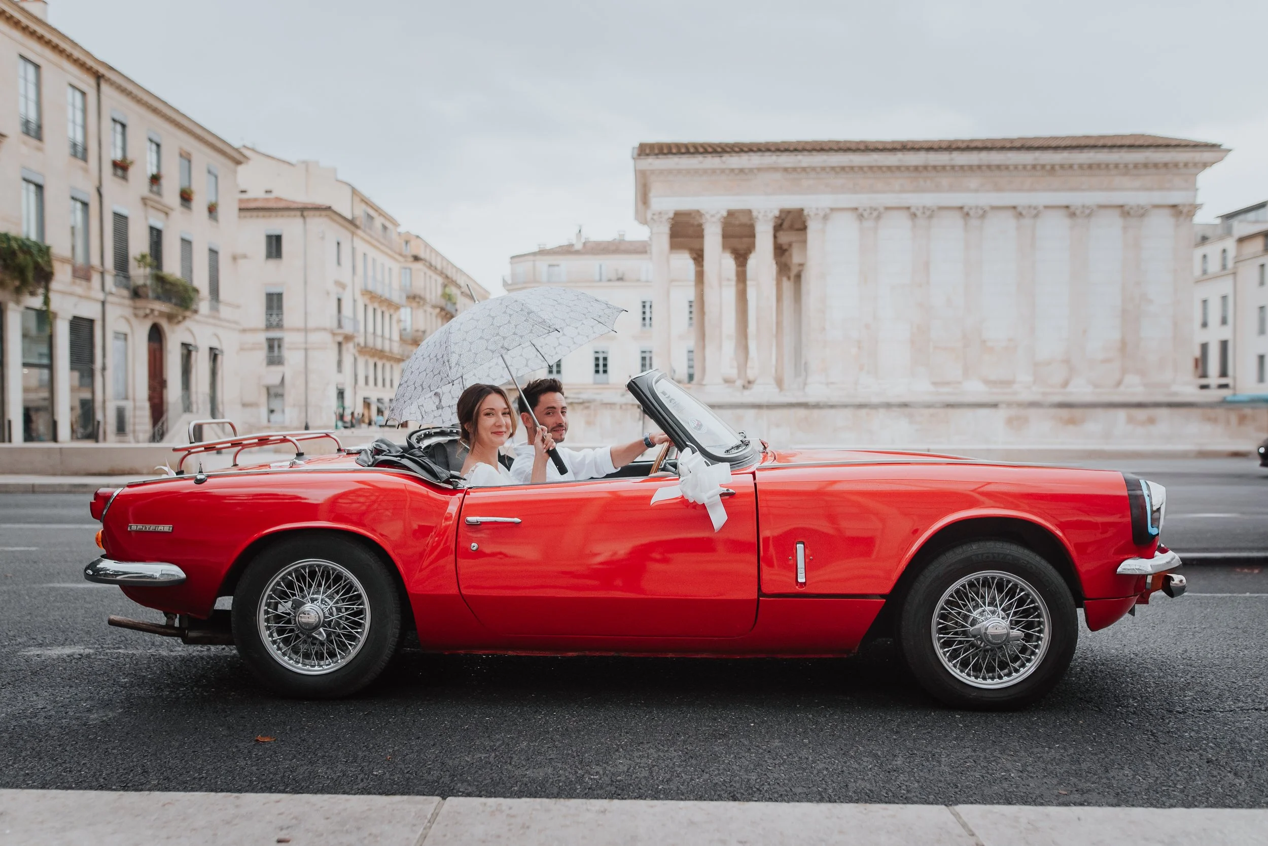 Un couple assis dans une voiture décapotable rouge avec un manège blanc devant l'Acropole d'Athènes, sous un parapluie, lors d'une journée nuageuse