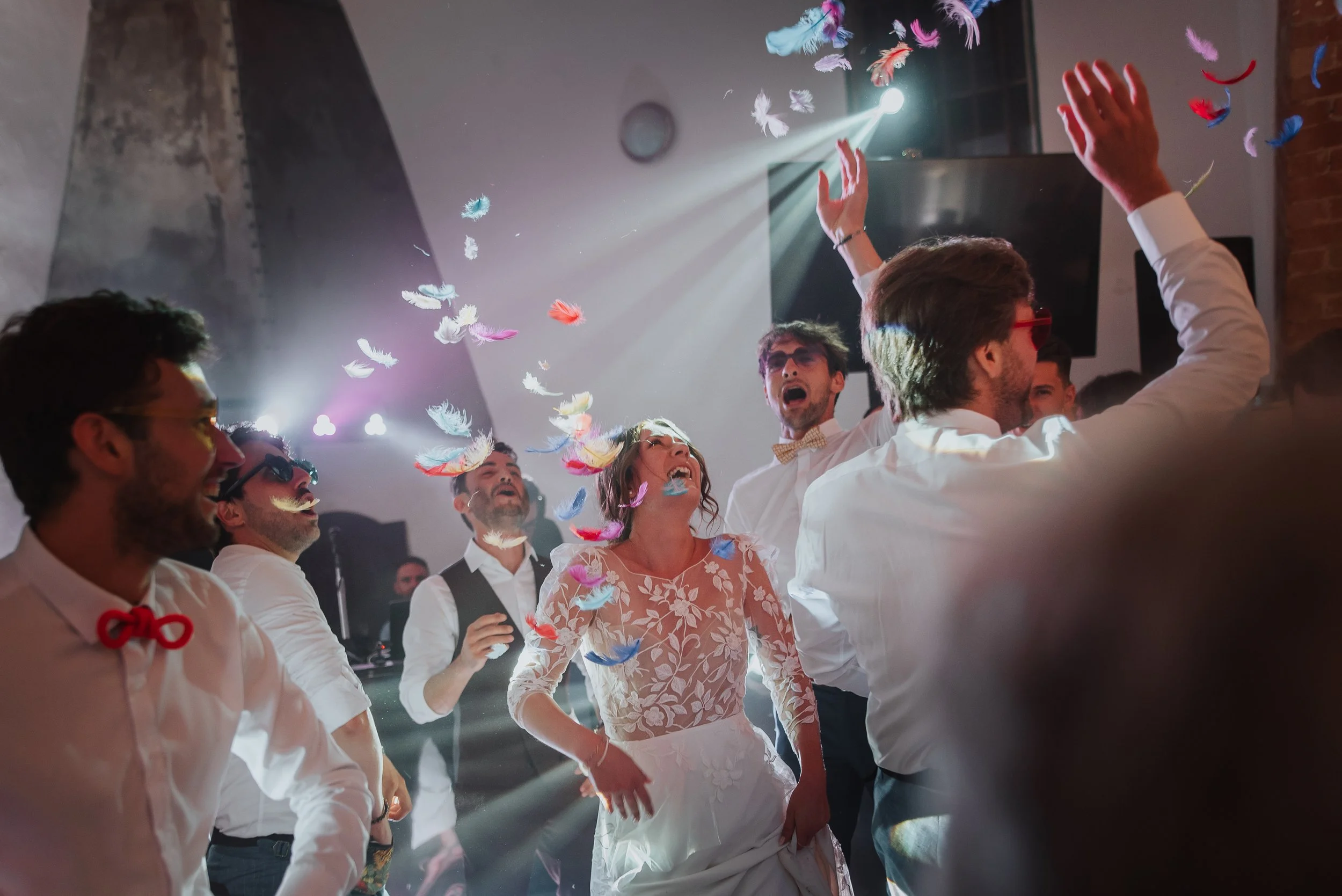 Groupe de personnes dansant et s'amusant lors d'une fête, avec des plumes colorées volant dans l'air, dans une ambiance festive avec effet de lumière.