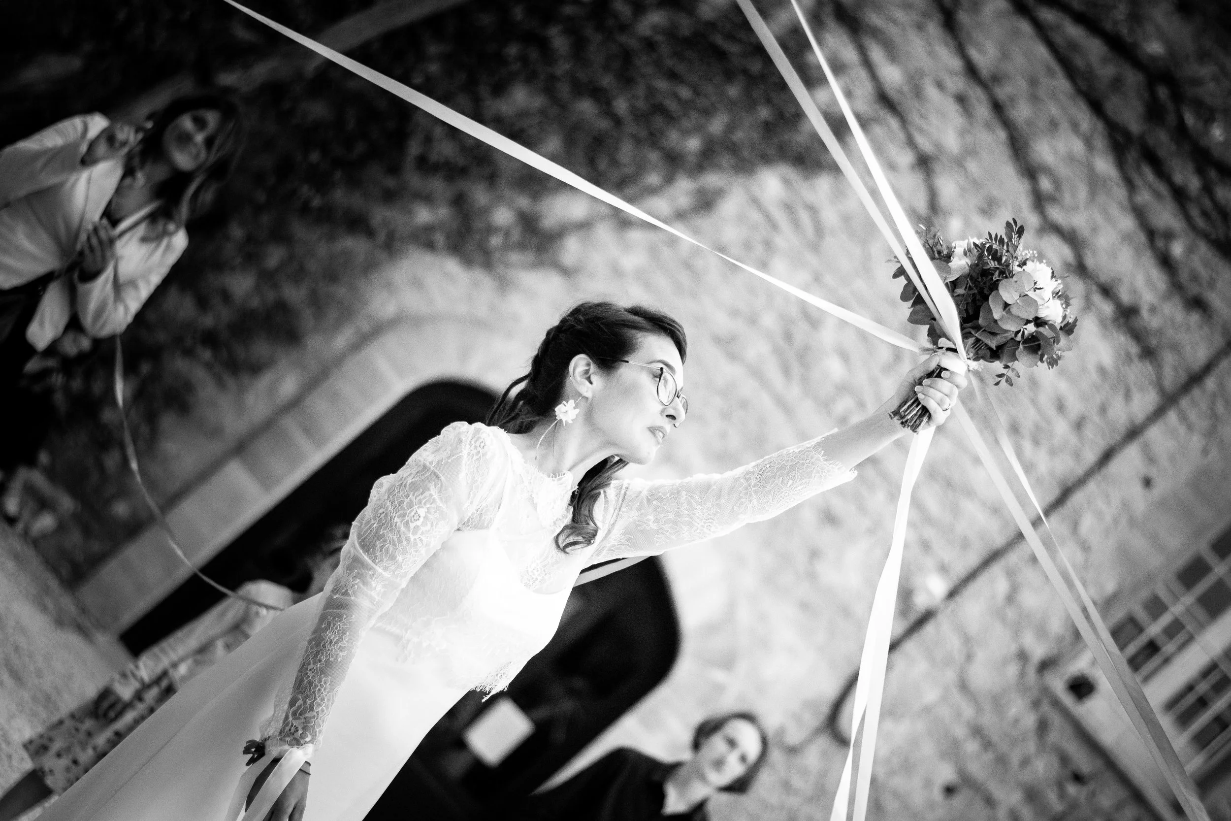 Femme en robe de mariée tenant un bouquet, lors d'une cérémonie dans une enceinte en pierre, avec des personnes en arrière-plan.