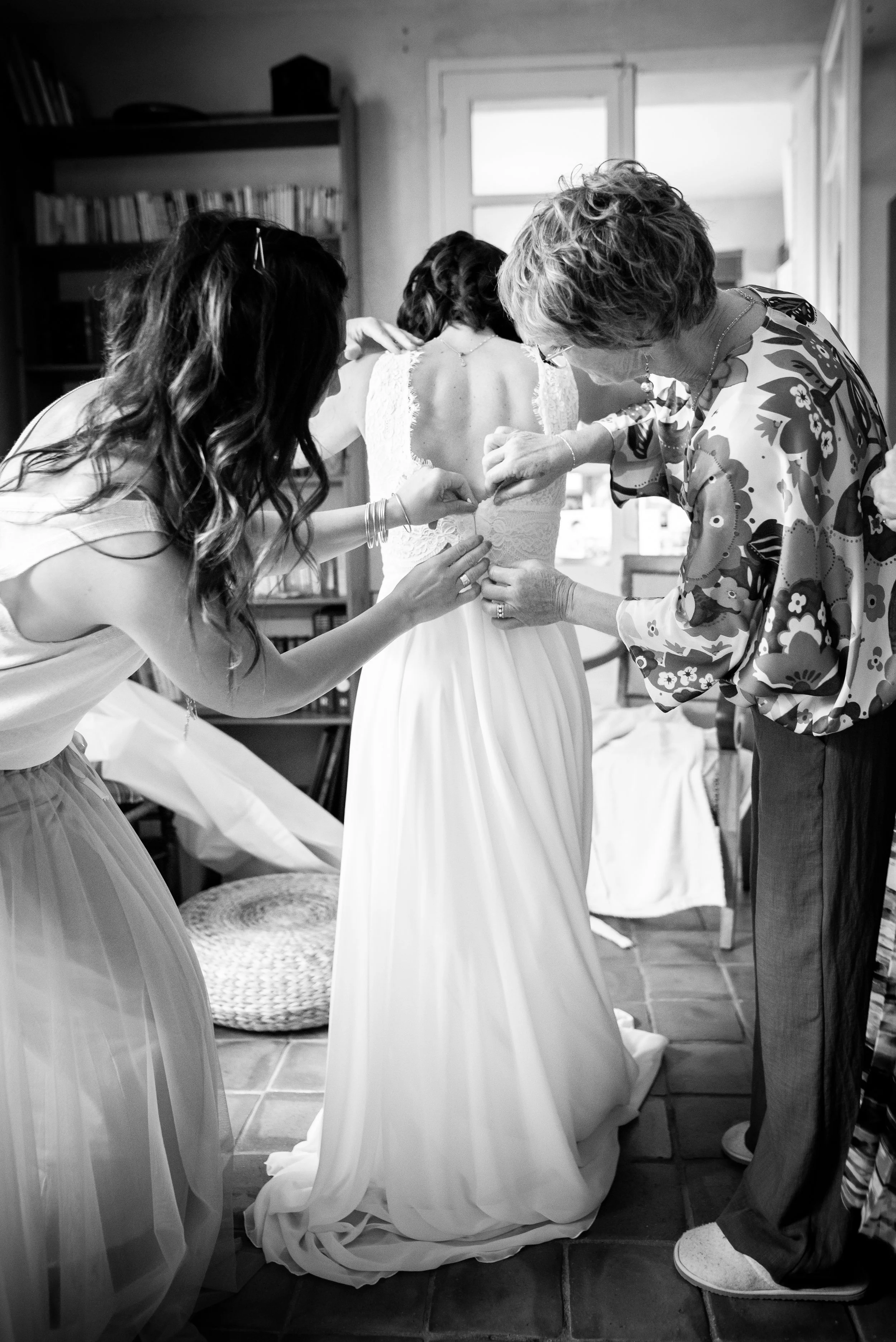 Une femme en robe de mariée est aidée à boutonner la robe, entourée de deux femmes lors de la préparation pour un mariage dans une pièce bien éclairée.